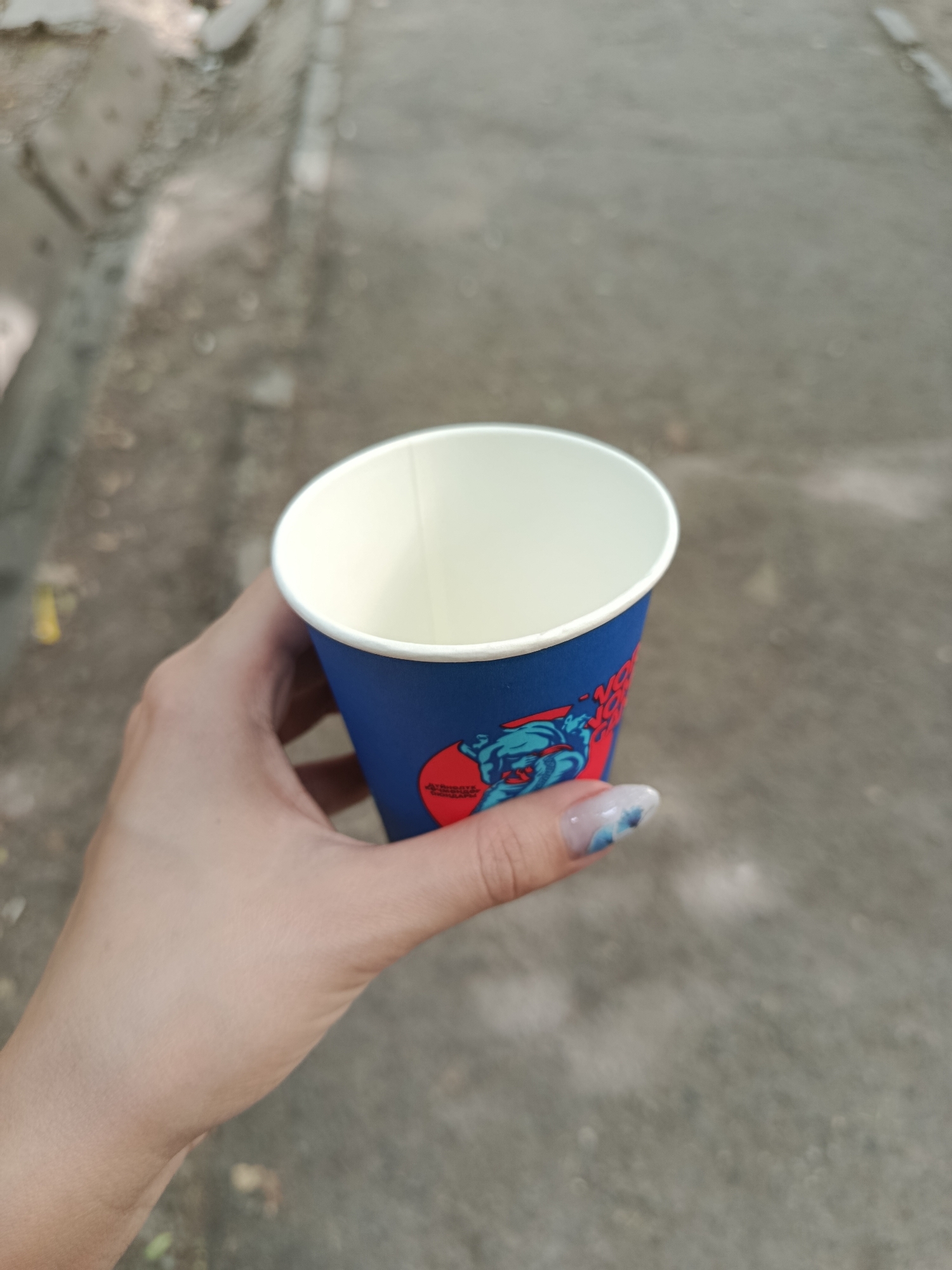 A person's hand holds a blue paper coffee cup with a red and blue design. The cup is empty and open at the top, revealing a white interior. The background is a blurred, gray paved surface, suggesting an outdoor urban environment. The person's fingernails are visible, decorated with a blue and white marble-like design. The scene appears to be outdoors, possibly in a park or on a sidewalk in Bishkek City, Kyrgyzstan.