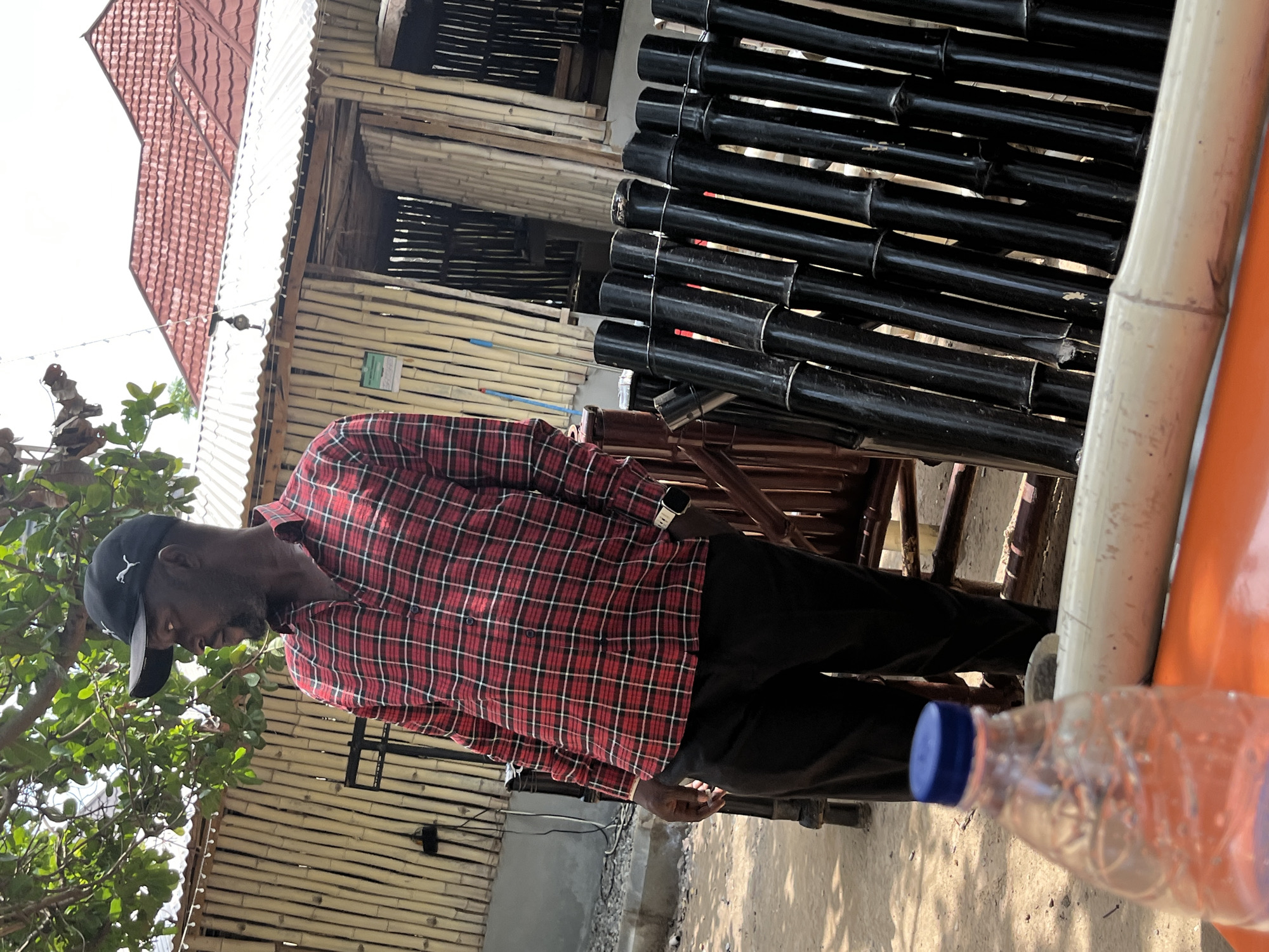 A man wearing a black baseball cap and a red and black plaid shirt stands outdoors. He is looking down towards the ground, with his head tilted. The man is also wearing black pants. He appears to be standing next to a structure made of bamboo poles and what looks like corrugated metal roofing. To his right, there is a clear plastic water bottle with a blue cap, lying on its side. The background shows more of the bamboo structure and some trees with green leaves. The sun appears to be shining, casting shadows on the ground, suggesting it is daytime. The overall setting appears to be informal, possibly a market or an outdoor seating area. There is no visible text in the image.