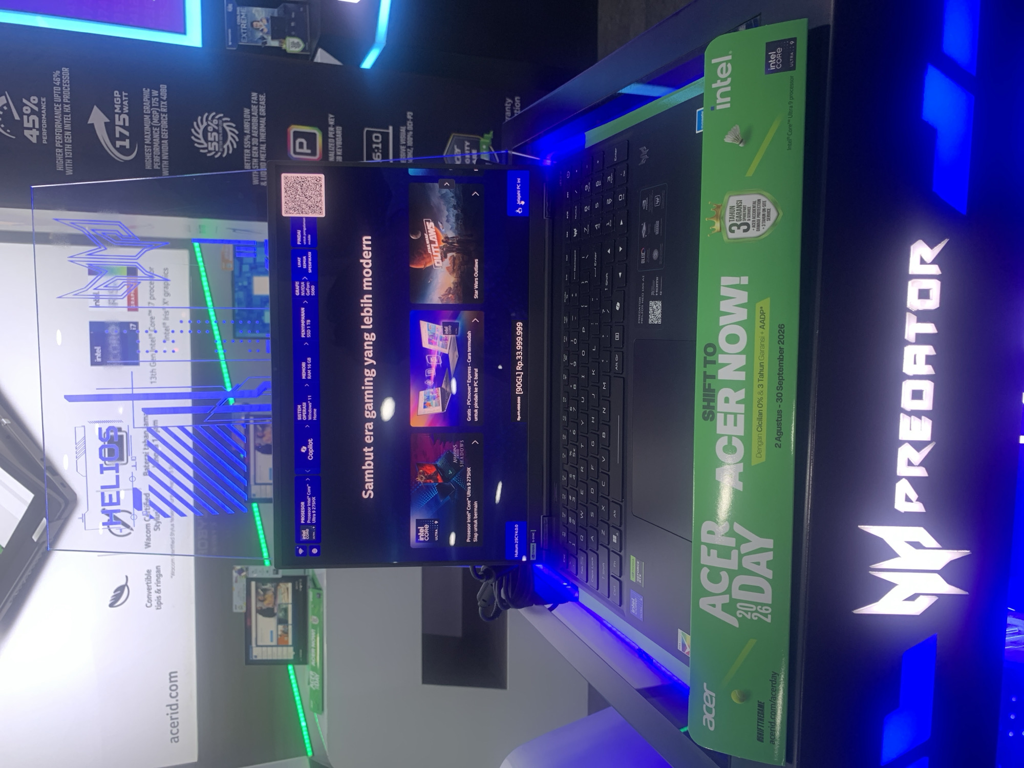 This is a photograph of a display booth for Acer Predator gaming laptops. The booth features a laptop with a prominent "Acer Predator" logo on the screen. The screen displays promotional material for gaming content, including game titles like "Assassin's Creed Shadows" and "Star Wars Outlaws." There are also specifications and benefits of the laptop highlighted on the screen, such as processor details and cooling system information. A green banner with "Acer 20-26 DAY" and promotional text is placed in front of the laptop. The overall setting appears to be an indoor retail or exhibition space, possibly a mall or electronics store. The lighting is dim with blue and green accent lights illuminating the display. There are no people visible in the image.