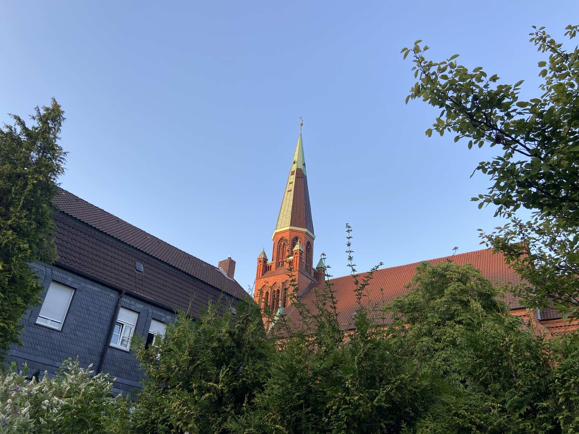 The image shows a view of a church spire and building, partially obscured by trees and other buildings. The sky is clear and blue. The church is made of red brick and has a tall, green spire topped with a cross. In the foreground, there is a building with a dark, tiled roof and grey, slate-like siding. Trees are scattered throughout the scene, with lush green foliage. The overall impression is of a calm, possibly late afternoon or early morning scene in a European city.