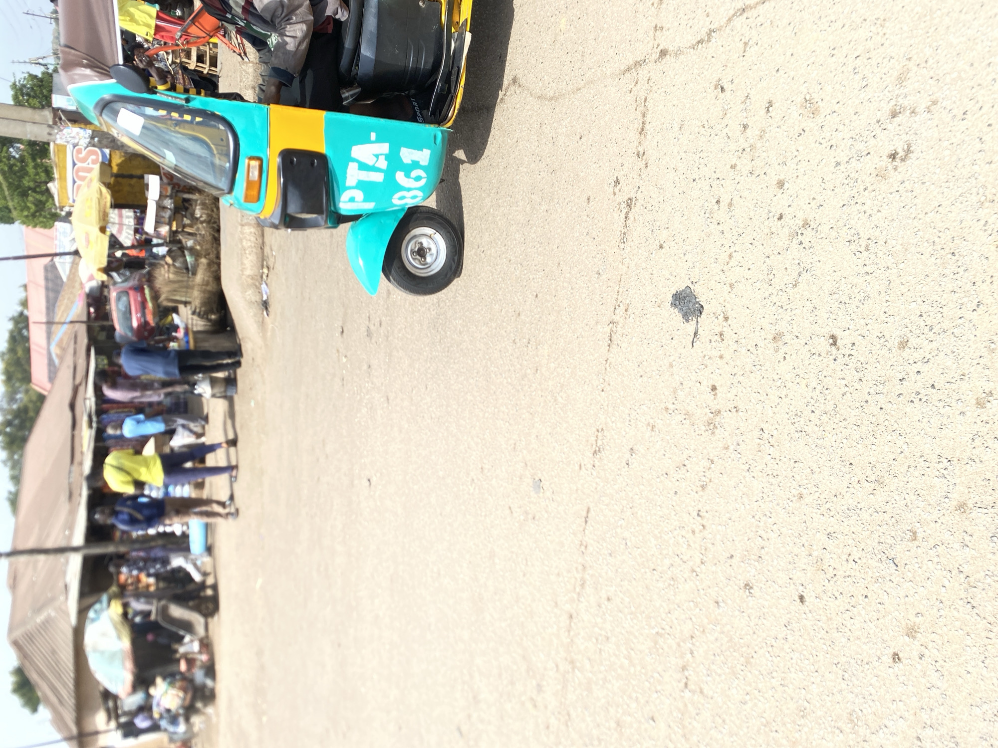 The image shows a three-wheeled vehicle (auto rickshaw/tuk-tuk) parked on a paved road in what appears to be a commercial area in Bukuru, Nigeria. The vehicle is primarily blue and yellow with "PTA-861" printed on its side. The street is lined with stalls or shops, with several people standing and walking around. The lighting is bright, suggesting daytime.