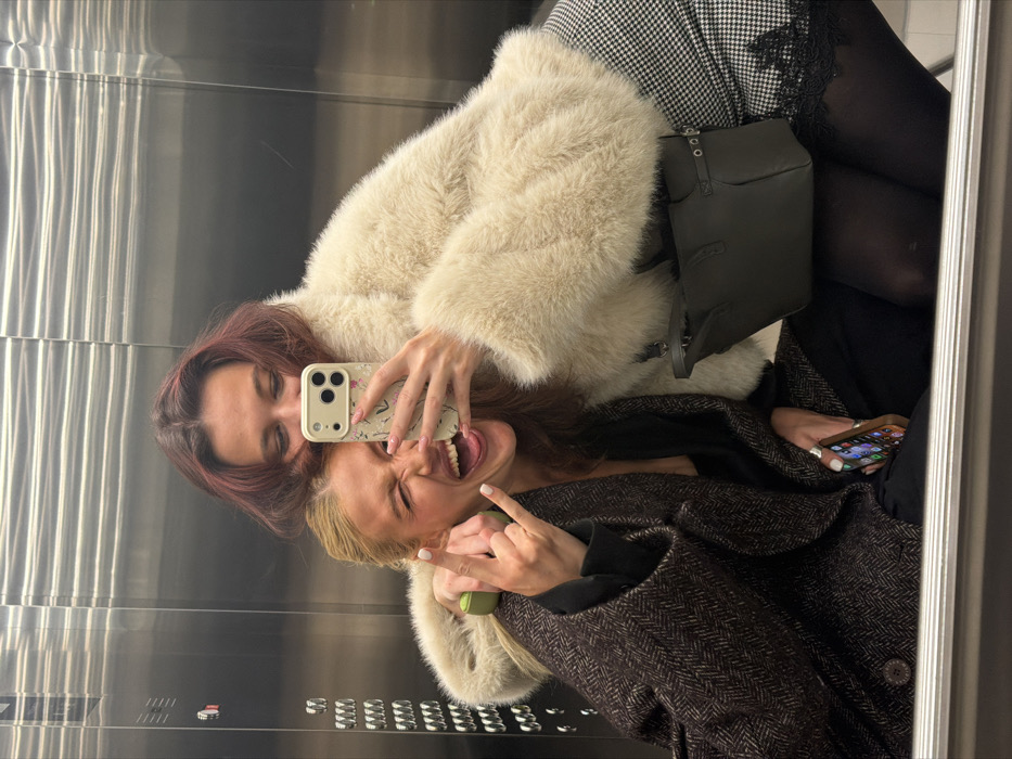 Two women are reflected in an elevator mirror. The woman on the left wears a beige fur coat, and her hair appears maroon. She holds a phone with a floral case, taking a selfie. The woman on the right, in a brown coat, sticks out her tongue and holds up two fingers in a playful gesture. A dark green bag is visible, along with a glimpse of patterned fabric and black hosiery. The second woman is also holding her own phone.