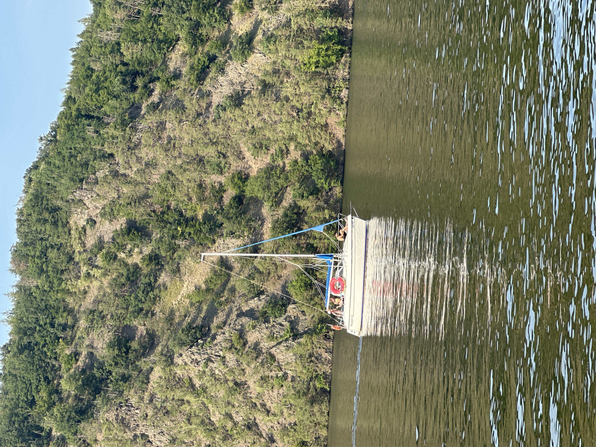 The image shows a sailboat on a body of water, with a lush green, rocky hillside in the background. The sailboat has a white hull and a blue sail. Several people are visible on the deck of the sailboat, appearing to be enjoying a leisurely ride. One person is standing, and others are seated. A red life preserver is attached to the side of the boat. The water is calm, with gentle ripples reflecting the sunlight and the sailboat. The hillside is densely covered with trees and shrubs, with exposed rocks adding texture to the terrain. The sky is clear and light blue, suggesting a sunny day. The scene appears serene and peaceful, likely taking place in the late morning or early afternoon, given the bright lighting. There is some text on the side of the sailboat, but it is too small and blurry to be legible.