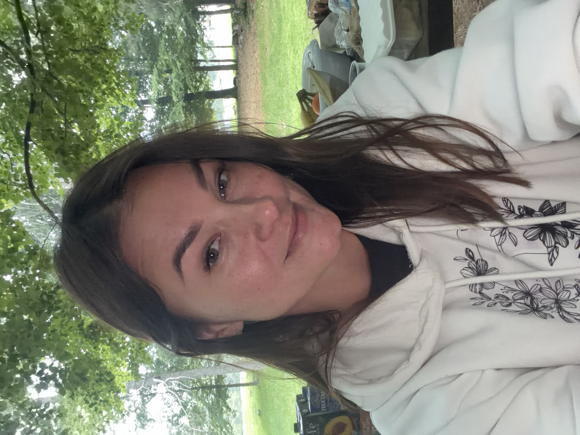 A young woman is captured in a selfie-style shot, looking directly at the camera with a soft, pleasant expression and a slight smile. She has long, dark brown hair and brown eyes. She is wearing a white hooded sweatshirt adorned with a black floral pattern featuring outlines of flowers and leaves.
The setting is outdoors, characterized by abundant green foliage from trees that fill the background, suggesting a forest, park, or natural recreational area. Patches of green grass are also visible. Behind the woman, on what appears to be an outdoor table, several items are scattered. These include plastic containers, bottles, and cardboard juice boxes. Specifically, one box clearly displays the brand name "FRUCTAL" and the flavor "Peach." Other items on the table include what looks like bananas and possibly an orange, indicating a snack or meal laid out.
The image appears to be taken during the daytime, with natural light, possibly under an overcast sky or in a shaded area due to the soft lighting and lack of harsh shadows. The overall atmosphere is relaxed and casual. The location context provided is Mali Bostur, Montenegro, consistent with the natural, outdoor environment depicted. The image is rotated 90 degrees counter-clockwise from a standard portrait orientation.