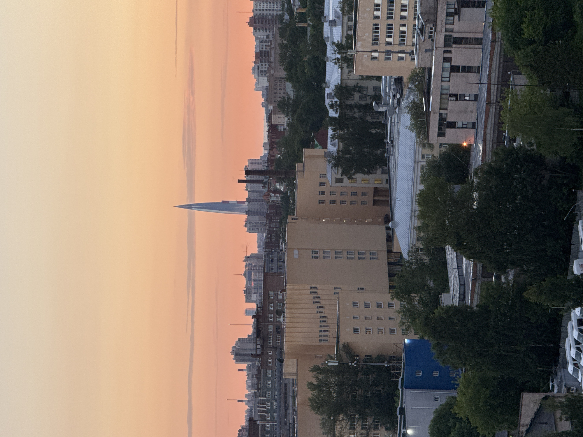 The image depicts a cityscape during what appears to be sunset, characterized by an orange and pink gradient in the sky. In the distance, a tall, slender skyscraper with a pointed top dominates the skyline. Numerous buildings of varying heights and architectural styles are visible, ranging from multi-story residential or office blocks to industrial-looking structures. Trees and foliage are interspersed throughout the urban landscape, particularly in the foreground and mid-ground. Several vehicles, including white vans, are parked in what looks like a lot or street in the lower portion of the image. There are no visible people in the image. The scene is urban, with a mix of modern and older architecture. The lighting suggests it is either early morning or late evening. The location is identified as Sinyavino, Russia.