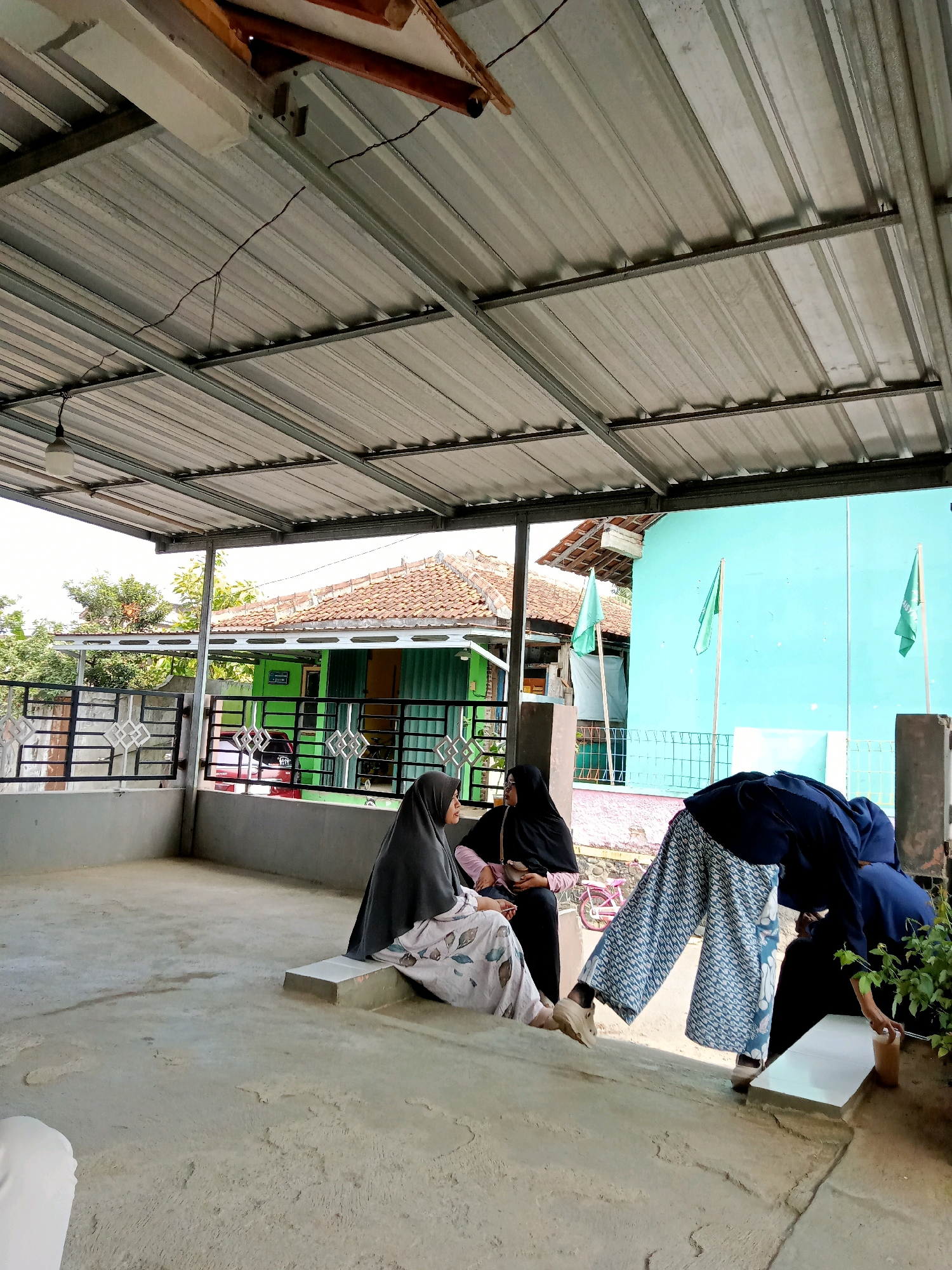 The image depicts an outdoor scene under a corrugated metal roof, likely in Gumiwang, Indonesia. In the foreground, three individuals are visible. Two women, dressed in hijabs, are seated on a concrete step on the left. One is wearing a gray hijab and a patterned skirt, while the other wears a black hijab. To the right, another individual, also in dark attire, is bent over, tending to something near a small plant in a pot. A patterned blue and white garment is draped over a bench in the mid-ground.
Beyond the covered area, a bright turquoise building with two green flags stands out. A red car is parked near a green building with a tiled roof in the background, suggesting a residential or community area. A black metal fence with a decorative pattern separates the seating area from the view of the street. The ground is a rough, concrete surface. The overall impression is of a casual, daytime gathering in a semi-public space.