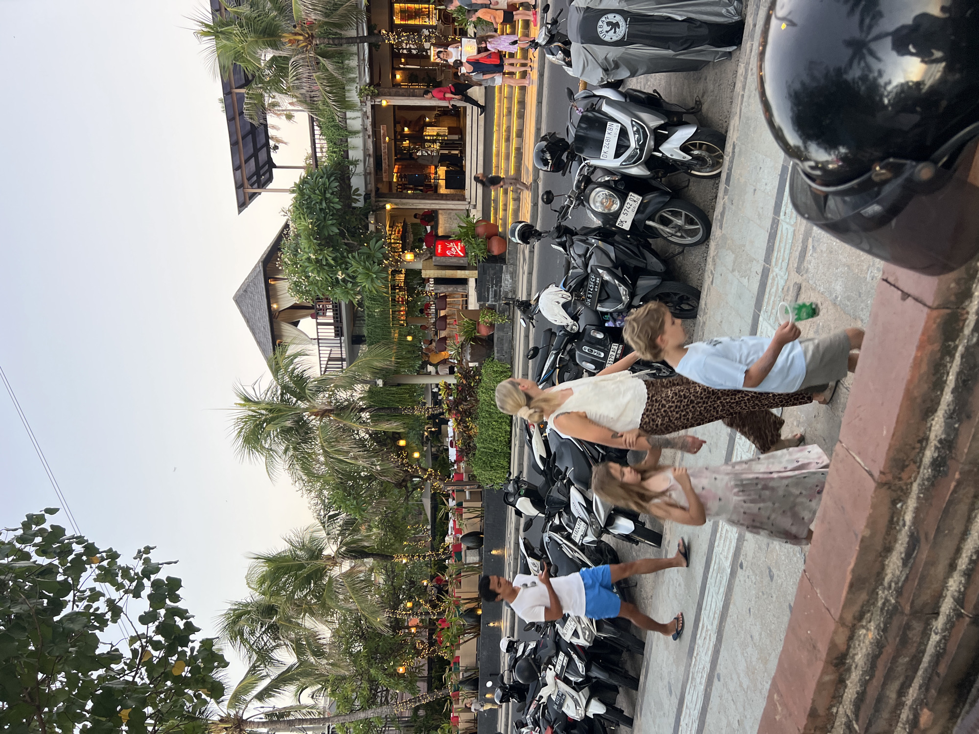 In Kuta, Indonesia, on a paved pedestrian walkway, a family walks along a row of parked scooters. A man in blue shorts and a white t-shirt walks ahead, holding a phone. Beside him, a woman with blonde hair tied back in a ponytail wears a white tank top and a leopard-print skirt, holding the hand of a young girl in a light-colored, patterned dress. A young boy in a light blue t-shirt and shorts walks slightly ahead of them. Behind the people, many scooters are parked, some covered with gray tarps. Palm trees and tropical foliage line the background, with strings of yellow lights illuminating a building with an open-air restaurant and bar. The sky is a soft, hazy gradient of pale blue and pink, suggesting late afternoon or early evening.