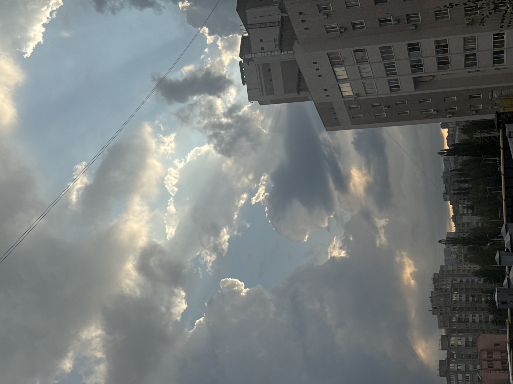 The image depicts a view of apartment buildings in Ufa, Russia, under a cloudy sky with shafts of sunlight breaking through. The buildings appear to be of Soviet-era construction, characterized by their uniform facades and numerous windows. In the foreground, several multi-story apartment blocks are visible, with trees interspersed between them. The sky is filled with dramatic clouds, some dark and heavy, others illuminated by the sun, creating a sense of depth and movement. Two thin power lines stretch across the upper portion of the frame. The overall impression is a typical urban landscape during a transitional weather period.