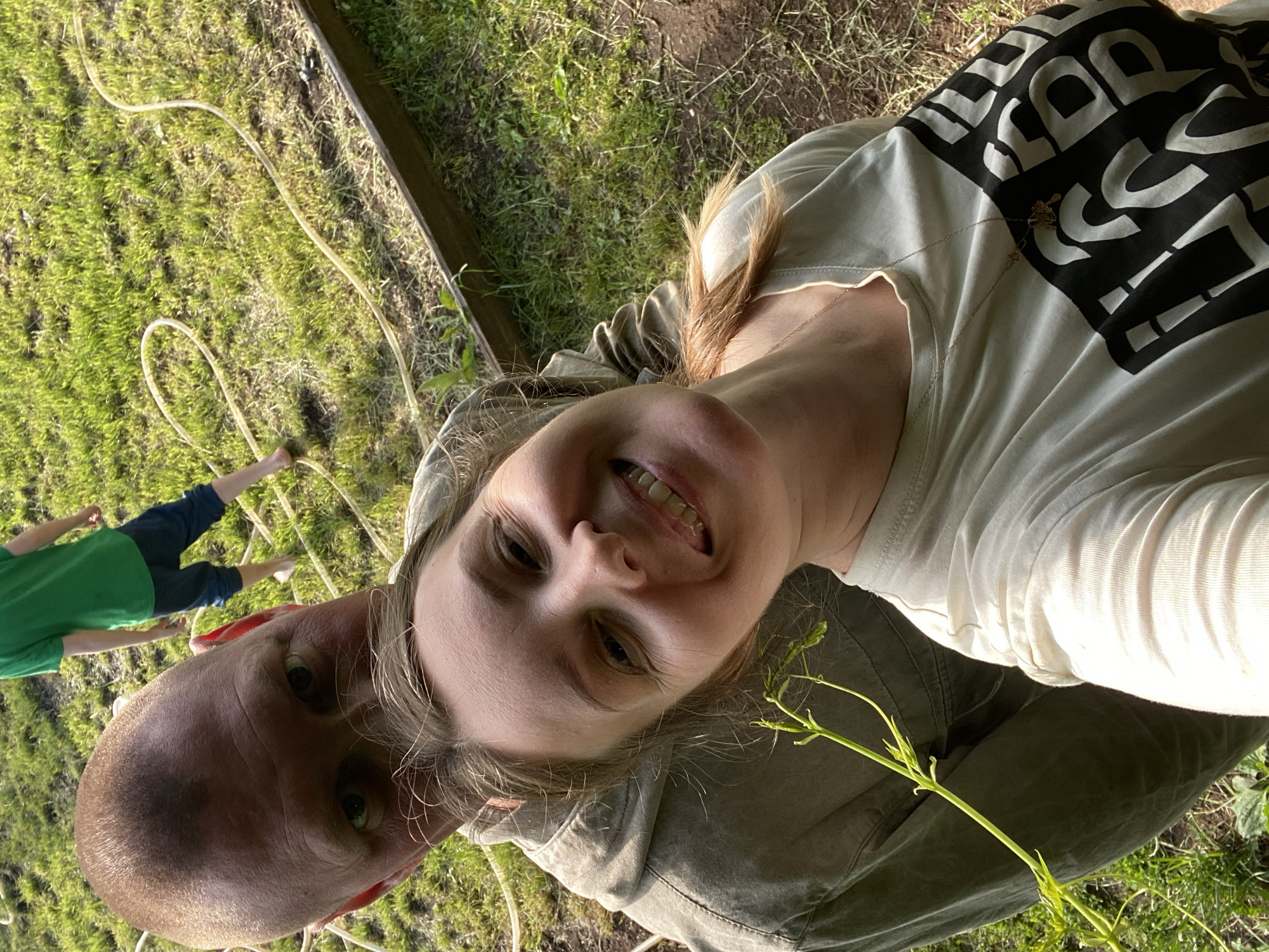 A selfie-style photograph captures two adults and a child in an outdoor, sunny garden or backyard setting, likely in Chaikovsky, Russia. The image is rotated, with the subjects appearing sideways.
In the foreground, a woman smiles brightly at the camera, holding the phone to take the picture. She has light brown hair and is wearing a light-colored t-shirt with a prominent black and white graphic design of stylized block letters across the chest, which are inverted and unreadable. A thin gold chain necklace is also visible. She appears to be wearing olive or khaki-colored pants or a jacket. A small green vine with leaves is near her shoulder.
Behind her and slightly to the left, a man with a shaved head is visible, looking towards the camera with a neutral expression. His face is partly obscured by the woman's head.
In the background, on the left side of the frame, a child is partially visible. The child wears a green t-shirt and blue shorts or pants, with bare feet. They are standing on green grass, near a beige garden hose that snakes across the lawn. The child appears to be interacting with the hose.
The setting features vibrant green grass and raised garden beds or planters with dark soil and some sparse vegetation. Bright sunlight illuminates the scene, suggesting a daytime, possibly late afternoon, period. The overall mood conveyed by the smiling woman is cheerful and relaxed.