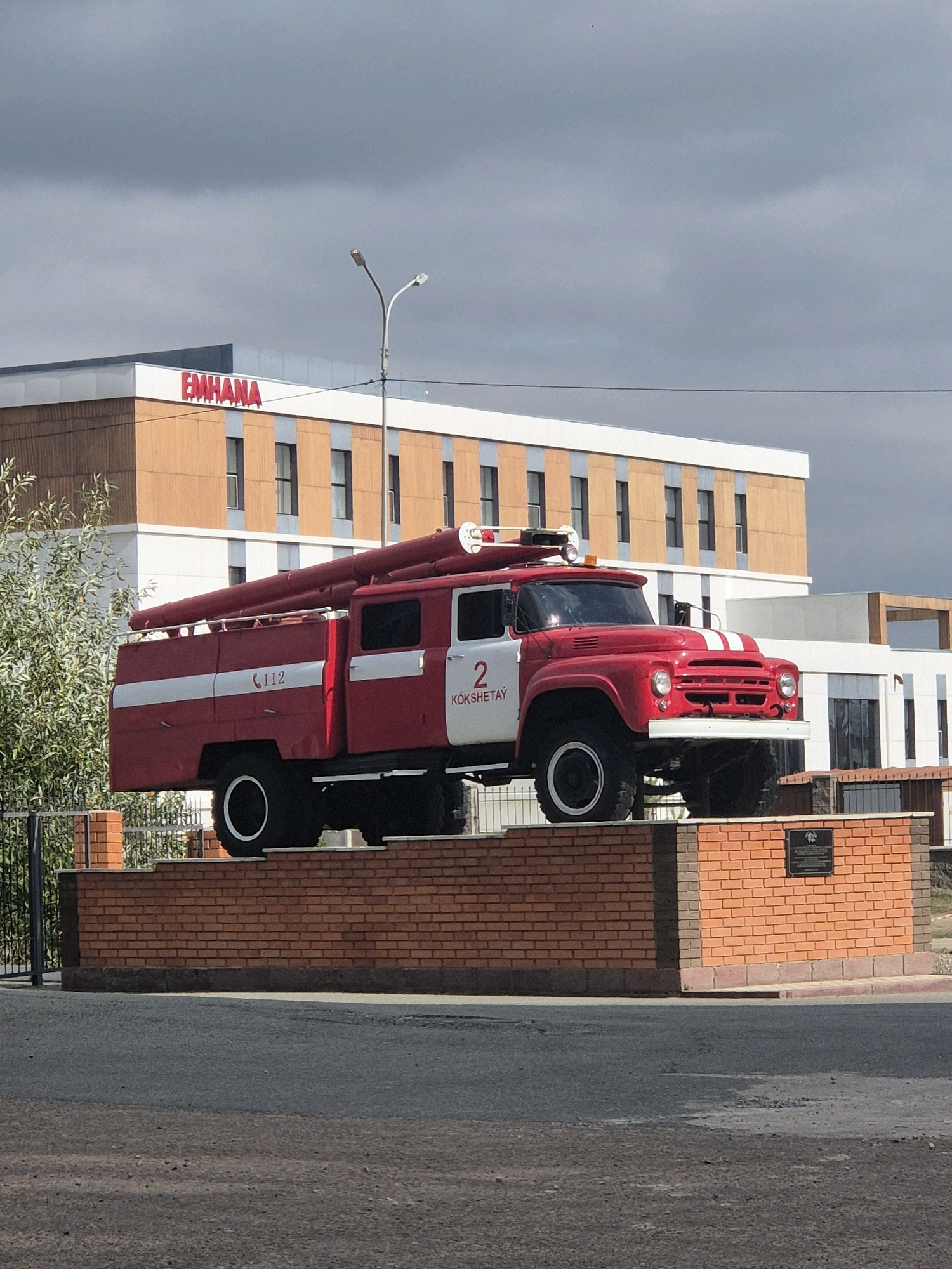 An exterior medium shot captures a weathered red and white fire truck monument situated on a brick pedestal in Kokshetau, Kazakhstan. The fire truck, a vintage model with a long hose bed, is positioned in the foreground, facing left. Behind the monument, a modern building with an "EMHANA" sign in red lettering is visible. The sky above is overcast with dark clouds. The scene is set outdoors, with a paved road in the lower portion of the image and a metal fence and foliage to the left of the fire truck. A lamppost stands behind the truck.