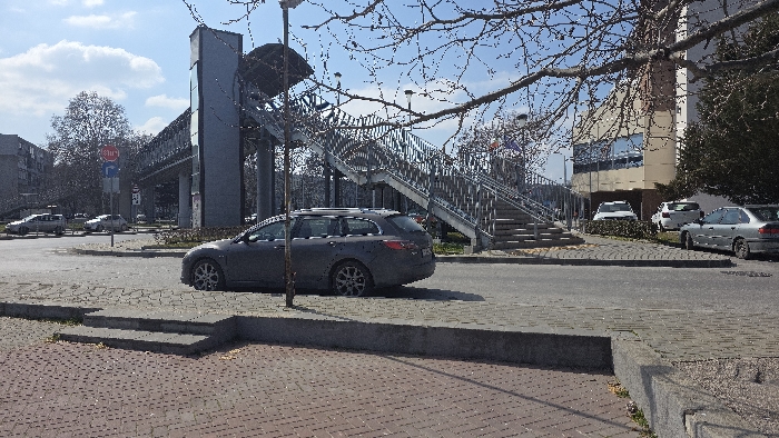 The image presents an urban scene in Varna, Bulgaria, specifically depicting the pedestrian overpass on Vladislav Varnenchik Blvd, likely in the vicinity of the Central Bus Station or Varna Mall area. The environment is under clear blue skies with scattered white cumulus clouds, suggesting fair weather.
In the immediate foreground, a reddish-brown brick-patterned paved surface is visible, transitioning to a grey concrete curb and a set of three steps. A dark grey Mazda 6 station wagon (third generation, 2012-2016 model), equipped with roof rails, is parked on the street's edge, oriented towards the left, occupying a central position in the lower midground. A slender, bare-branched tree or utility pole stands directly to the left of the parked vehicle.
The midground features an asphalt road surface with faint lane markings. Dominating the central and right portions is a large, grey metallic pedestrian overpass structure, comprising multiple sections of stairs with handrails ascending over the roadway. Several streetlights are mounted on and around the overpass. To the left of the overpass, a vertical pole displays a red octagonal STOP sign above a blue circular directional sign.
In the background, on the far left, a multi-story residential apartment building with a flat roof is visible. To the right, a modern, multi-story building with a light-colored facade and numerous windows, possibly a commercial or office structure, stands behind the overpass. Several vehicles, including a white SUV, another white car, and an older silver sedan, are parked near this building. Deciduous trees, largely bare of foliage, are dispersed throughout the scene, particularly noticeable in the upper right corner and behind the apartment building. No individuals are discernible within the frame.