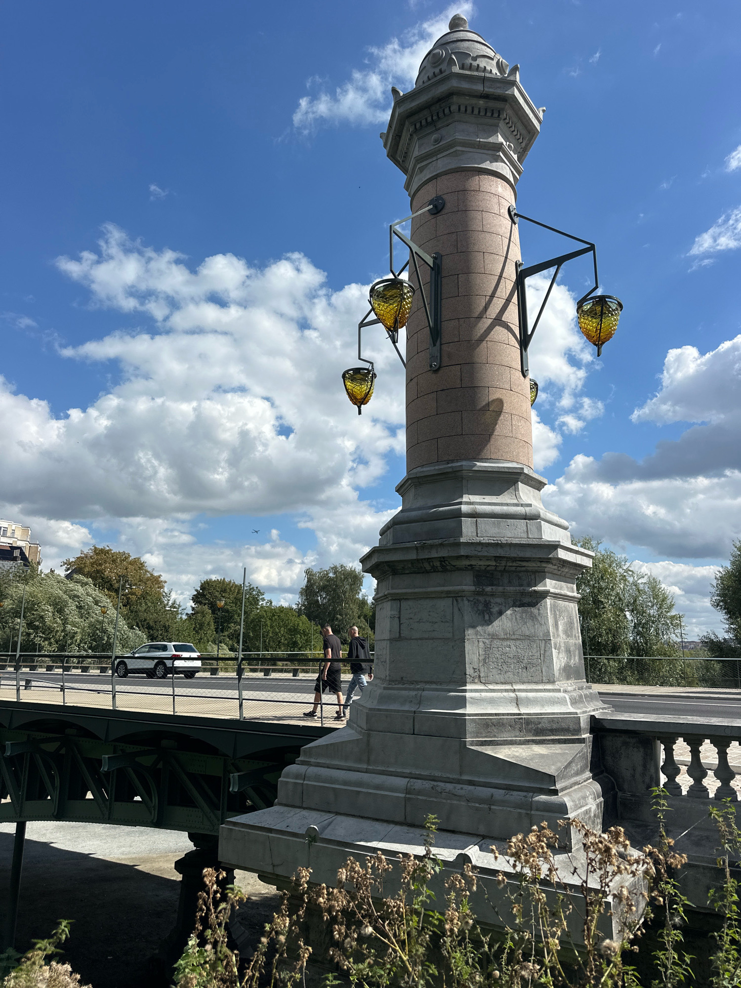 The image captures a scene on a bridge in Molenbeek-Saint-Jean, Belgium, on a sunny day with scattered clouds. In the foreground, a large, ornate lamppost stands prominently. This lamppost is made of stone and features multiple decorative elements, including four amber-colored glass lanterns attached to metal arms. The base of the lamppost is substantial, with layered stone construction.
To the left of the lamppost, a bridge structure is visible, with a white car driving on it and two people walking across. The bridge has a metal framework underneath and a railing along the side where the pedestrians are walking. Beyond the bridge, there is a backdrop of lush green trees and some buildings. In the sky, there are large, fluffy white clouds against a clear blue sky, and a small airplane is visible in the distance. The foreground also includes some dried vegetation in the lower part of the frame, suggesting a slightly unkempt natural area near the bridge. The overall impression is a well-lit, daytime scene on a functional urban bridge.