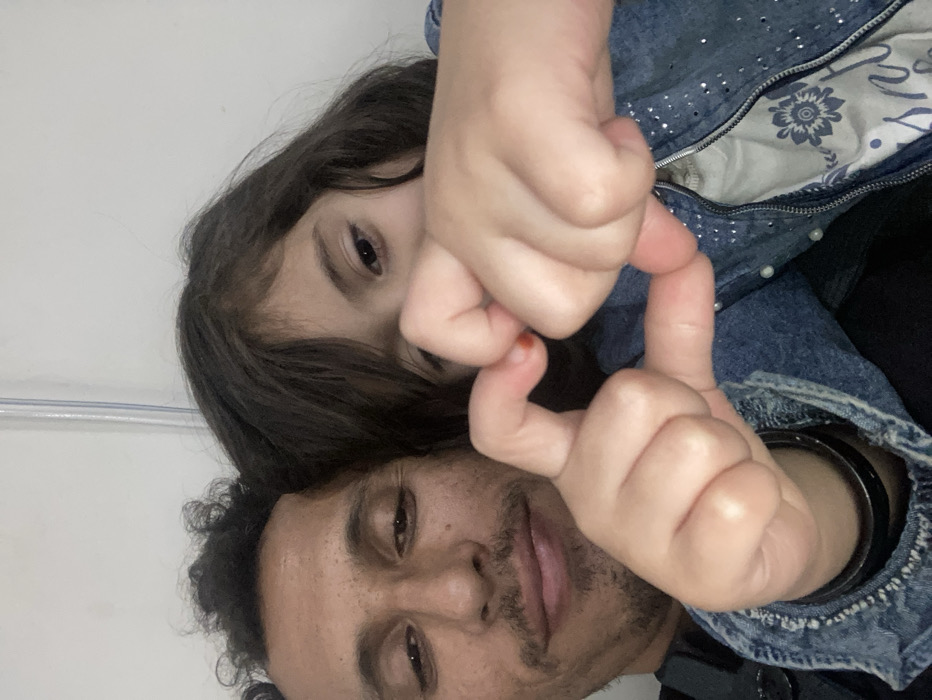 The image depicts a close-up portrait of two individuals, an adult male and a child, situated in Quetta City Tehsil, Pakistan. The adult male, on the left, has dark, curly hair, a mustache, and a short beard. He is wearing a dark shirt under a denim jacket, with a black bracelet on his left wrist. The child, positioned to the right, has dark, medium-length hair and is dressed in a light blue denim jacket with small, reflective embellishments over a light-colored top featuring a floral pattern. Both individuals have their hands in the foreground, forming a heart shape with their fingers. The adult male looks towards the camera with a slight smile, while the child's eyes are partially visible, also directed towards the camera. The background is a plain, light-colored wall with a faint vertical line, possibly a pipe or conduit, visible towards the top.