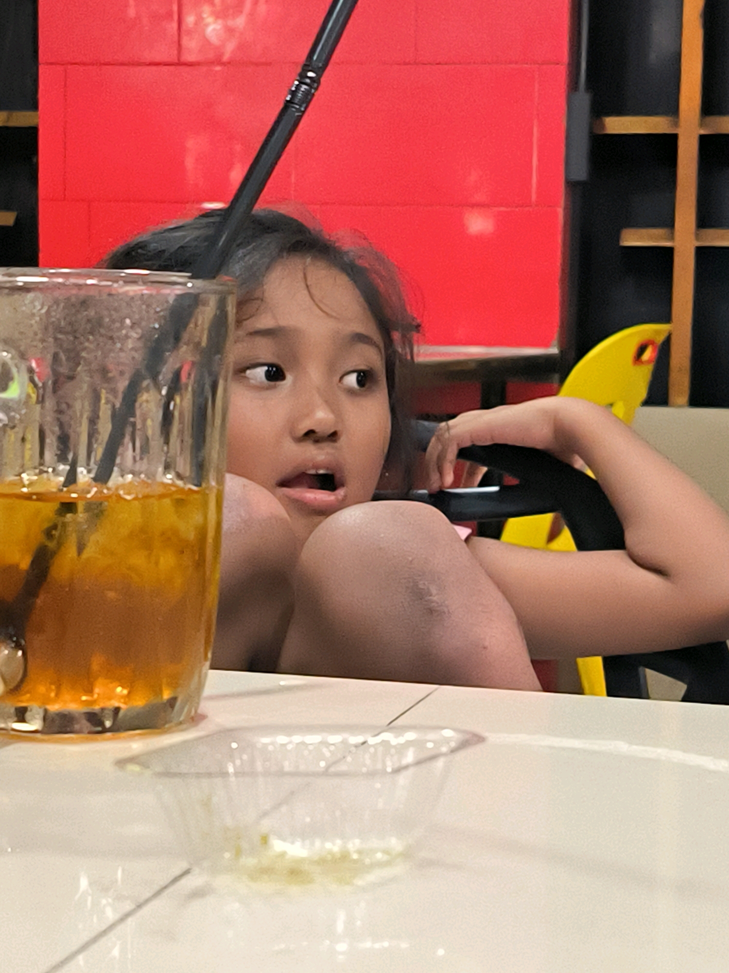 An Indonesian child with dark hair and eyes gazes off-camera to the right with their mouth open, as if in surprise or speaking. They are seated at a table with a glass of amber-colored liquid containing ice and a dark straw in the foreground. A black straw extends from the glass upwards towards the child's face. The background features a bright red wall made of rectangular blocks, a black shelving unit with yellow accents, and a yellow object resembling a part of a chair. The child is leaning their head on their arms, with their right arm resting on a black chair back. The setting appears to be a casual indoor environment, possibly a cafe or restaurant in Banda Aceh, Indonesia.