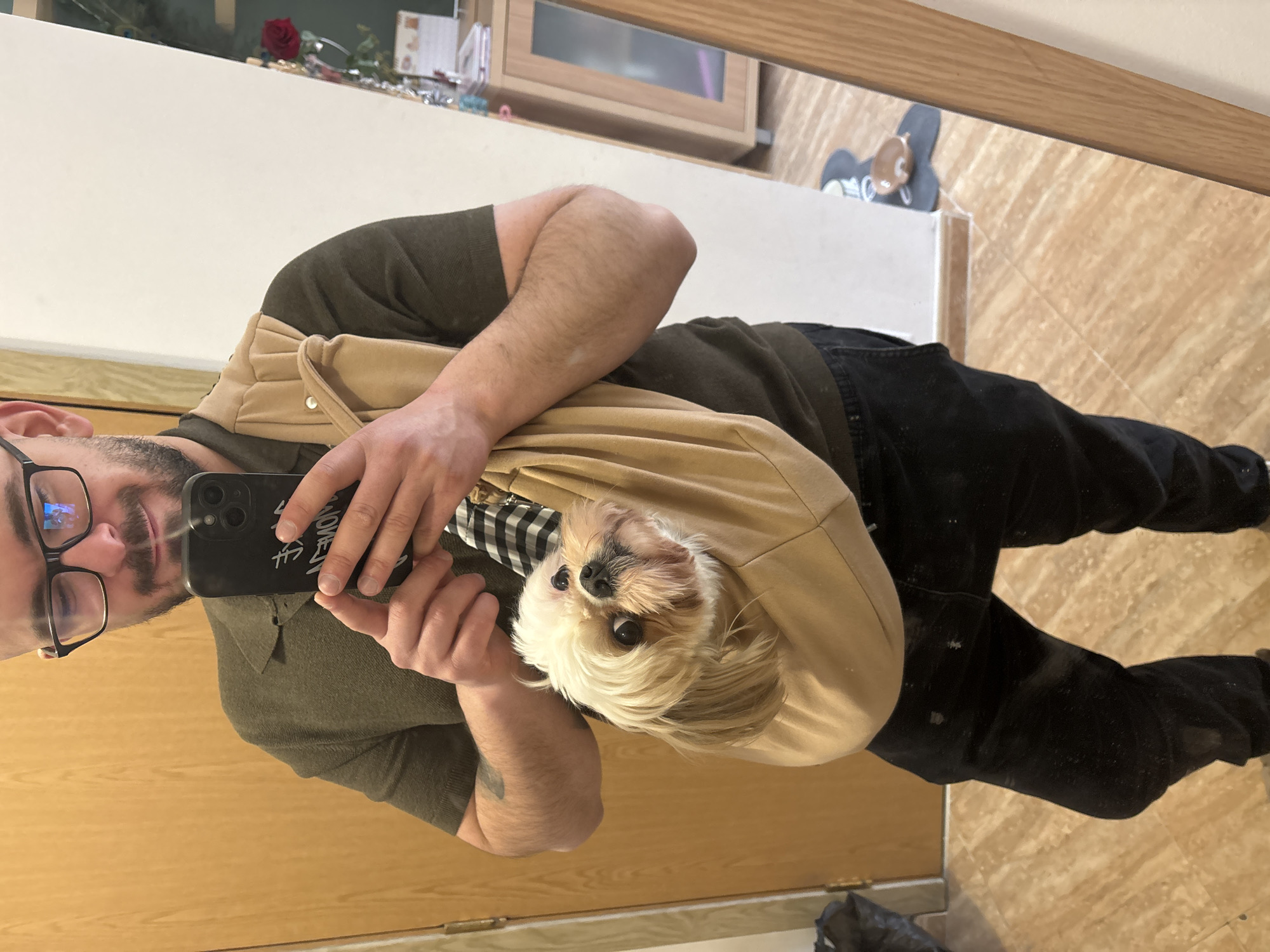 A man with glasses and a beard is holding a cell phone and taking a picture of himself with a small, white dog in a beige sling. The man is wearing a dark green shirt and black pants. The floor is tan and tiled. There is a mirror in the background, reflecting a rose in a vase, some toiletries, and a wooden cabinet. It is not possible to determine the exact location in Sofia, Bulgaria, from the image.