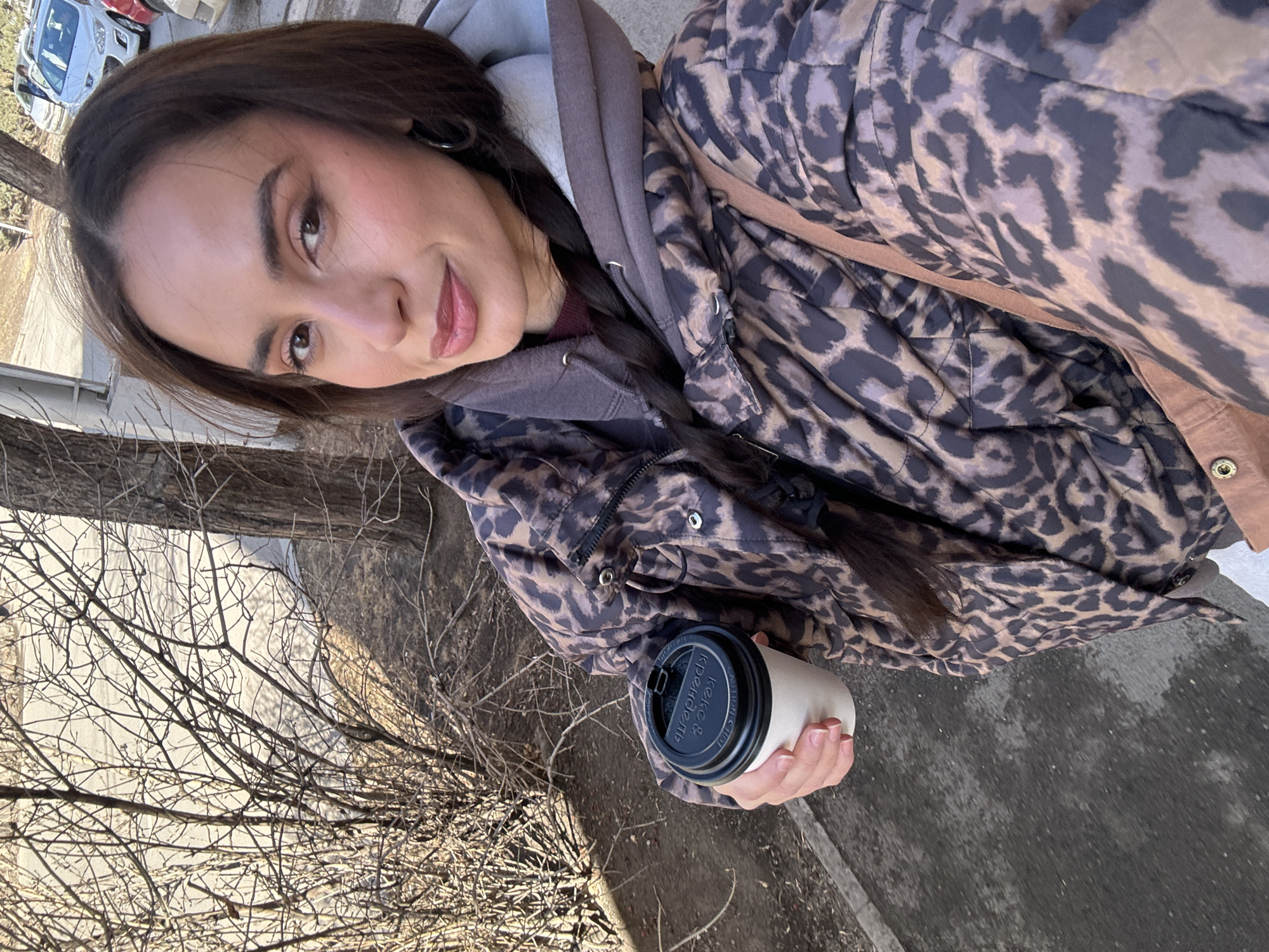 A woman is outdoors holding a paper cup of coffee with a black plastic lid. She is wearing a brown and black leopard-print jacket over a gray hooded sweatshirt. She is looking at the camera and smiling slightly. Behind her, a bare tree and gray pavement are visible. In the background, cars are parked on the street, suggesting an urban setting. While it is not possible to definitively state the location as Krasnoyarsk, Russia, without additional information, the style of buildings and vehicles is consistent with a modern urban environment and the lettering on the coffee lid is written in Russian.