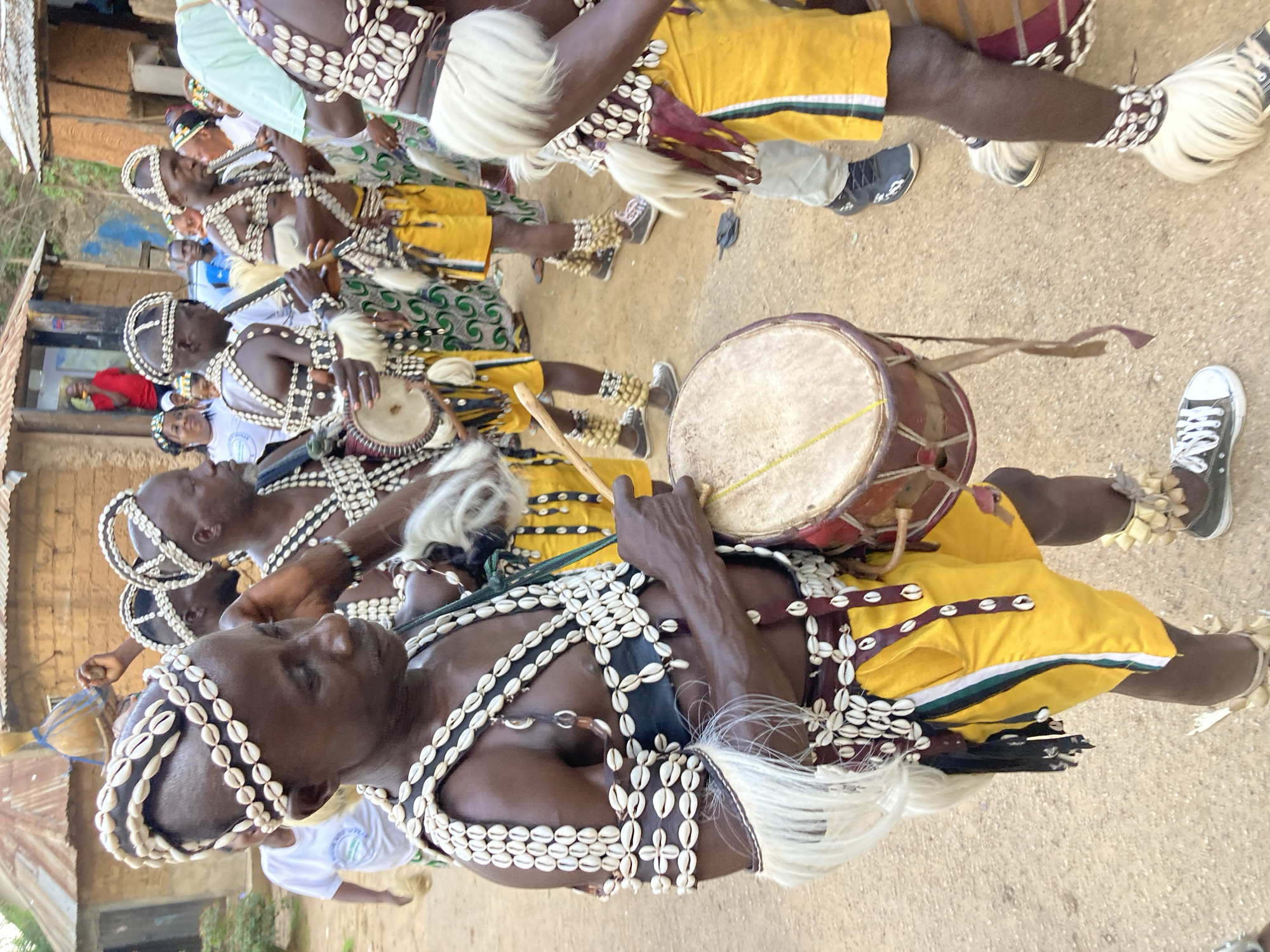 A group of men in traditional attire, adorned with cowrie shells and white fringes, are participating in a cultural event in the city of Jos, Nigeria. The man in the foreground, facing left, wears a headdress and a chest piece made of cowrie shells. He holds a drum stick and is about to strike a drum. Other men in the background also wear similar headdresses and chest adornments. They are dressed in yellow shorts with green and white stripes. Some men are holding musical instruments, possibly flutes. The environment appears to be an outdoor setting with buildings constructed of mud and corrugated iron roofs. The ground is sandy and dusty. A few women and children are visible in the background, observing the event.