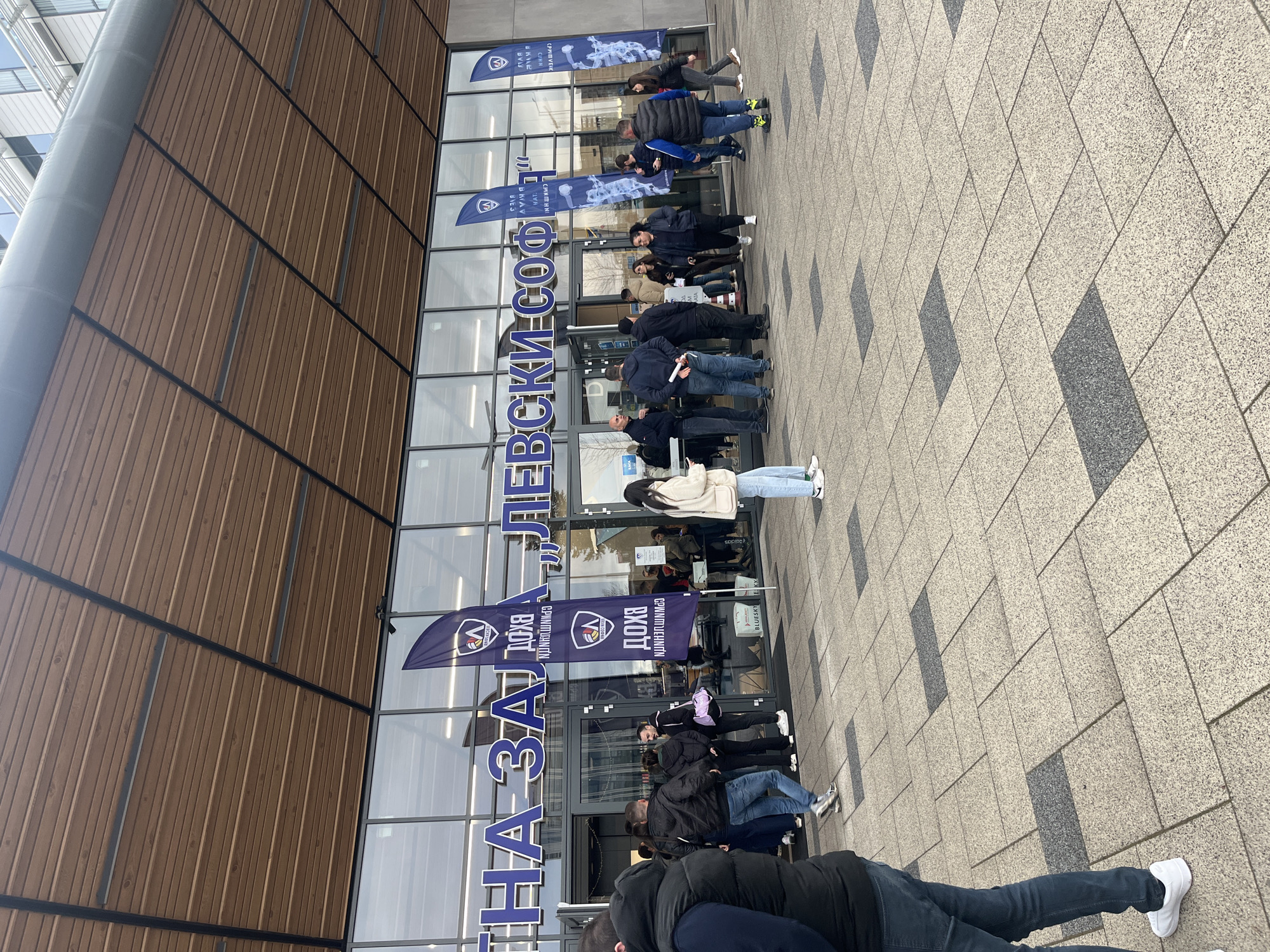 A medium, outdoor shot captures a line of people waiting outside a modern building in Sofia, Bulgaria. The building's facade is a combination of floor-to-ceiling glass windows and vertical wood paneling. Above the glass windows, large blue letters spelling "Левски Соф" (Levski Sofia) are visible. Banners with the Levski Sofia logo, a soccer team, are placed next to the entrance. People of various ages and in different clothing are standing in line. The foreground shows sections of the sidewalk, paved with stone tiles in a grey hue. The sky is overcast.