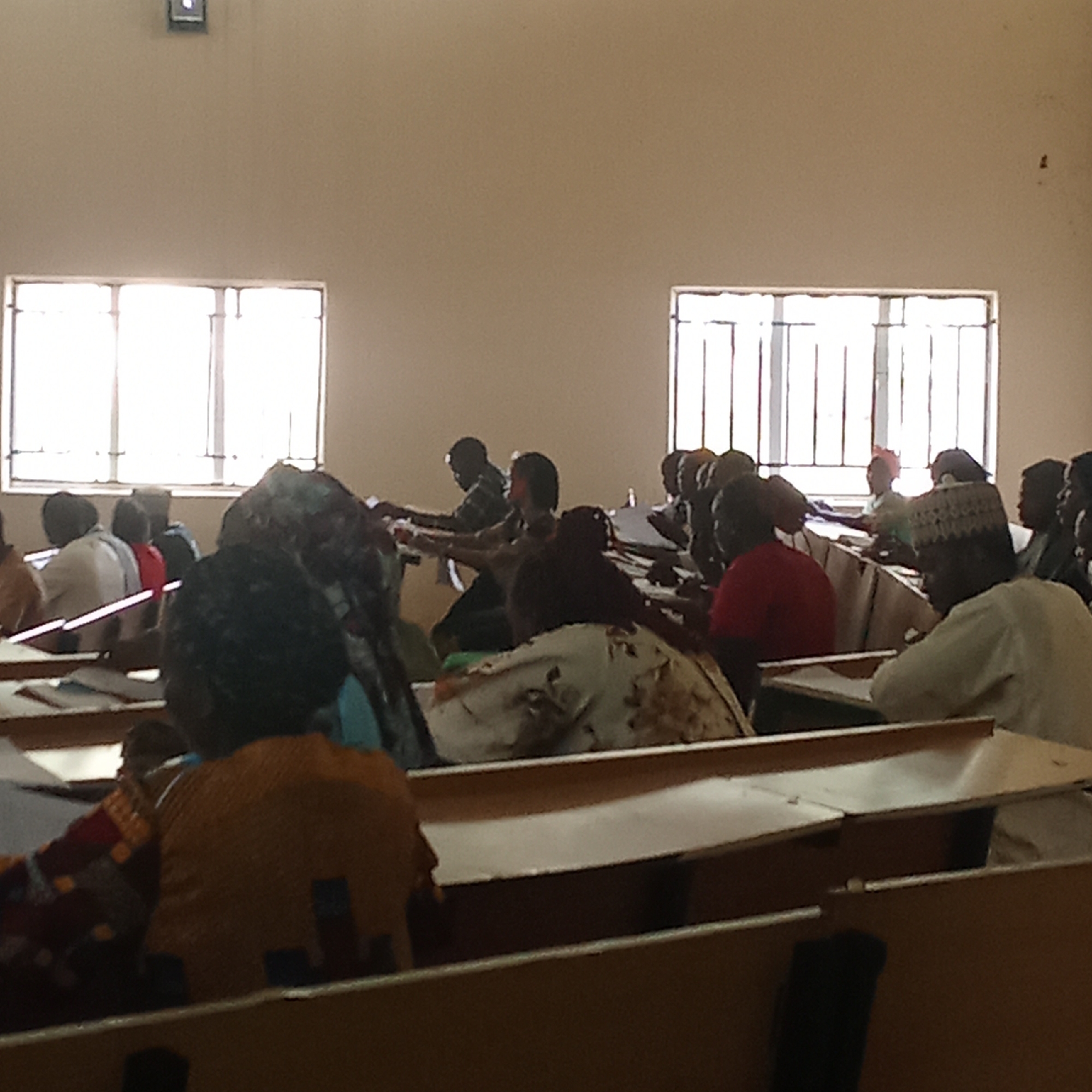The image depicts a classroom setting in Akwanga, Nigeria. Rows of desks are occupied by individuals, many of whom appear to be students. Most are seated and facing forward, presumably towards an instructor. Some are wearing traditional clothing. The room has two windows with metal bars, allowing natural light. The walls are painted in a light color. Some papers are visible on the desk surfaces.