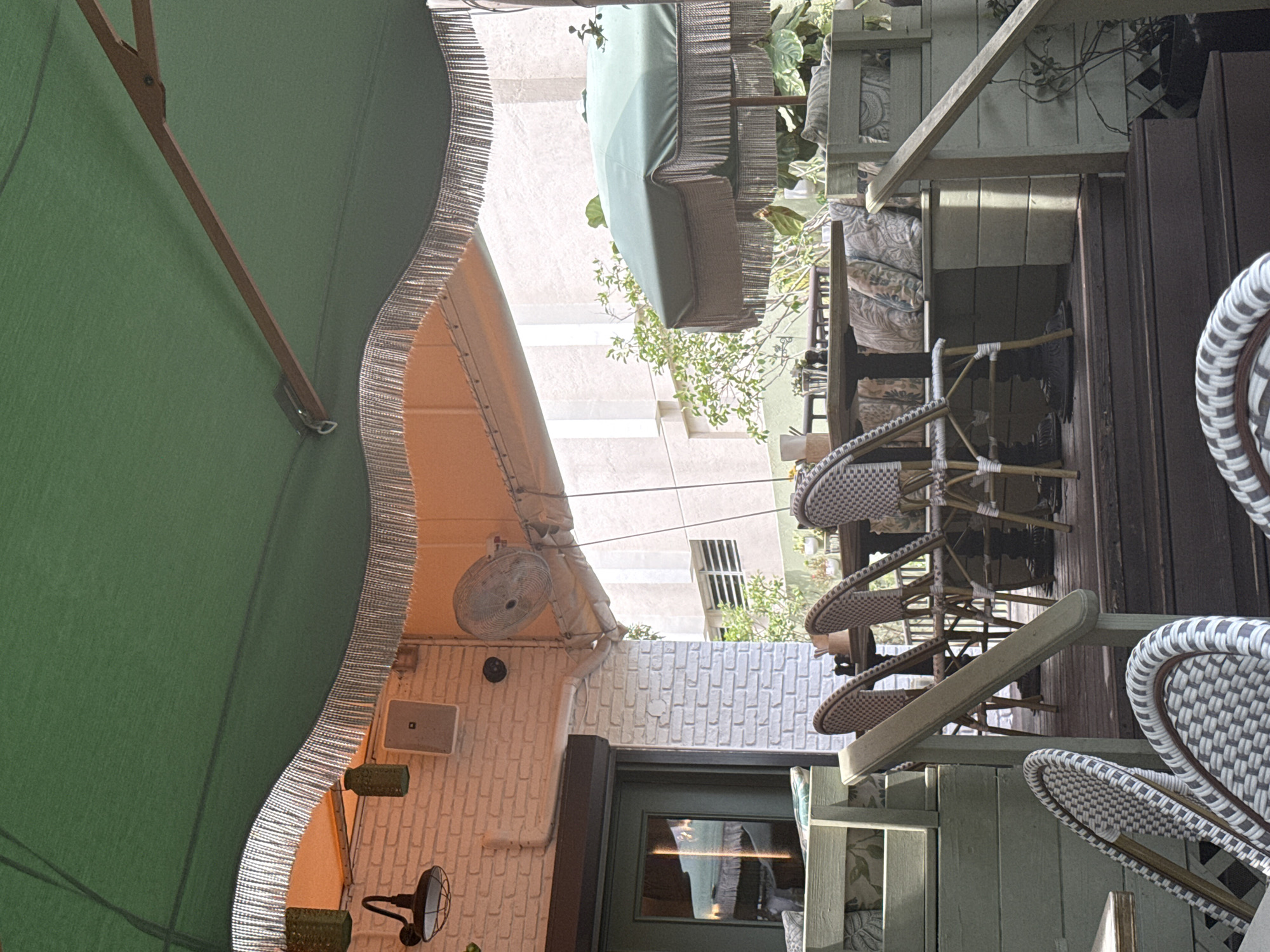 The image depicts an outdoor seating area, possibly a restaurant or cafe, located in Fort Lauderdale, United States of America. The scene includes various elements: a large green parasol partially covers the frame from the left, offering shade. Several wicker-style chairs and tables are arranged in a manner suggesting a dining area, with some chairs appearing stacked. A green awning is also visible. The architecture surrounding the seating area includes a white brick wall with mounted lighting and a ceiling fan. The presence of plants and trees indicates an outdoor setting. A doorway with a green frame is also discernible, leading indoors.