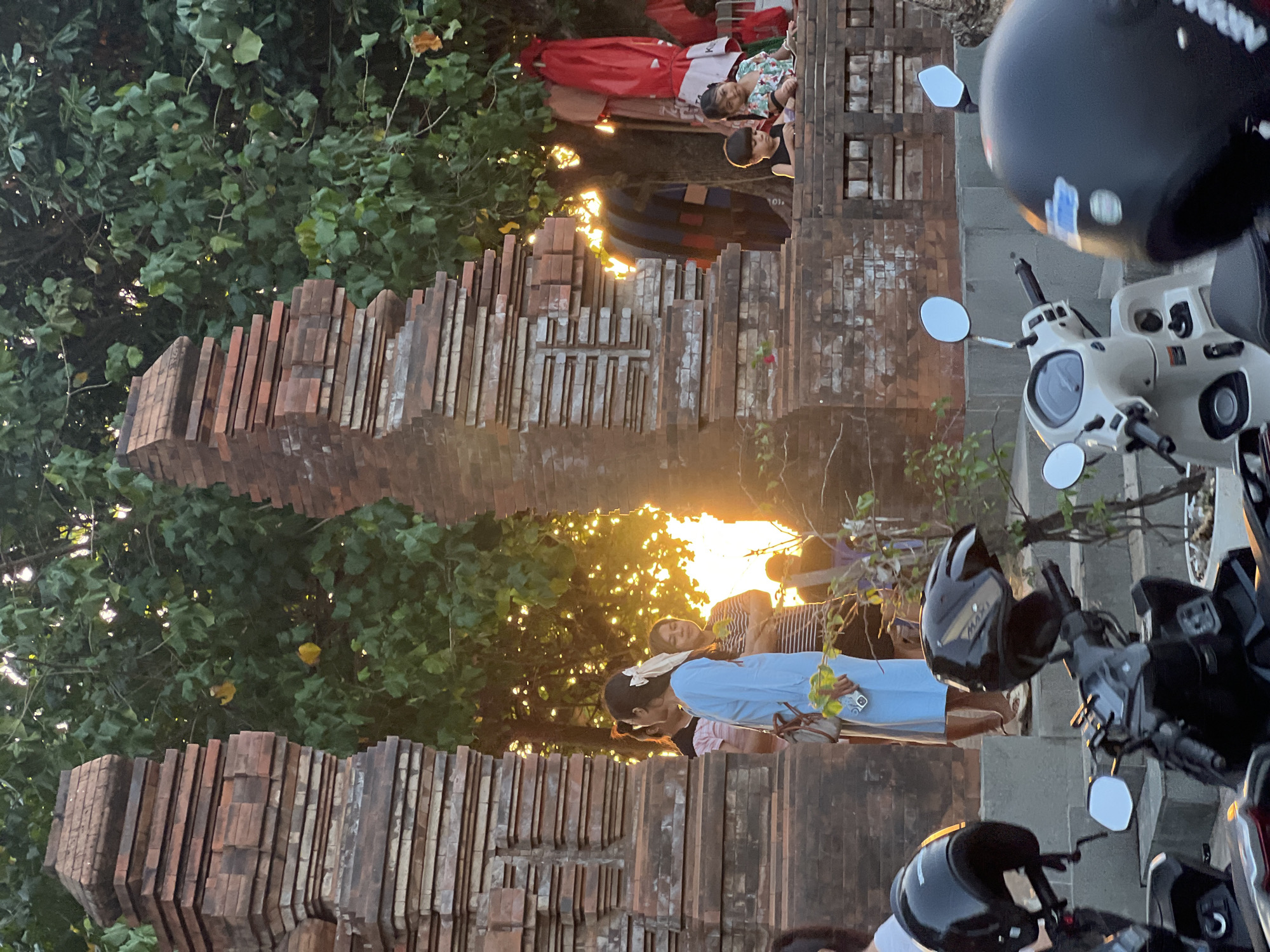 In Kuta, Indonesia, a street scene unfolds at sunset, illuminated by the warm glow of the setting sun filtering through lush green trees. Two imposing brick archways frame the view, their layered designs suggesting ancient architecture. Several people are visible, including a woman in a light blue dress with a bow in her hair, a woman in a striped shirt, and another individual partially obscured. In the background, two children are seated on a brick ledge. To the right, clothing and umbrellas are displayed, possibly from a nearby vendor. In the foreground, several parked motorcycles with black helmets evoke the everyday transportation of the area. The overall atmosphere is serene, capturing a moment of everyday life against a backdrop of cultural heritage.