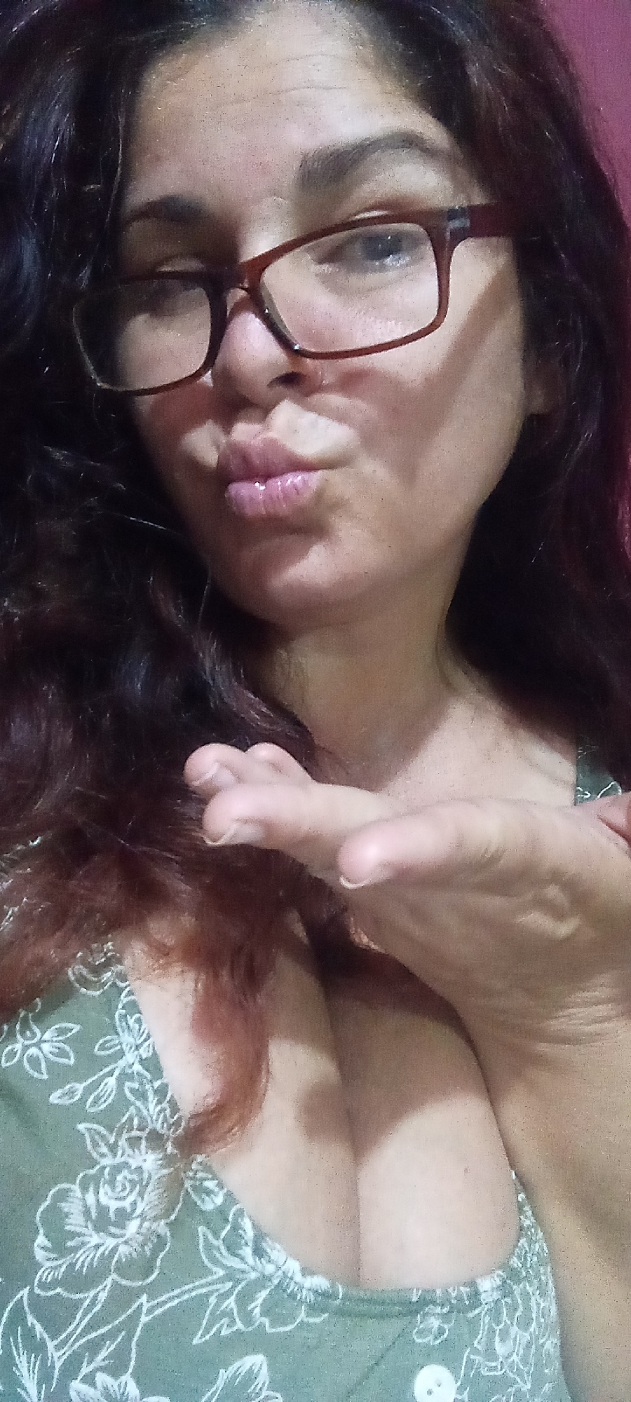 A woman with dark curly hair and glasses is posing for a selfie. She is wearing a green top with white floral patterns. Her lips are puckered as if blowing a kiss, and her hand is outstretched towards the camera. The background is blurry and indistinct, appearing to be a room with a pinkish-red wall.