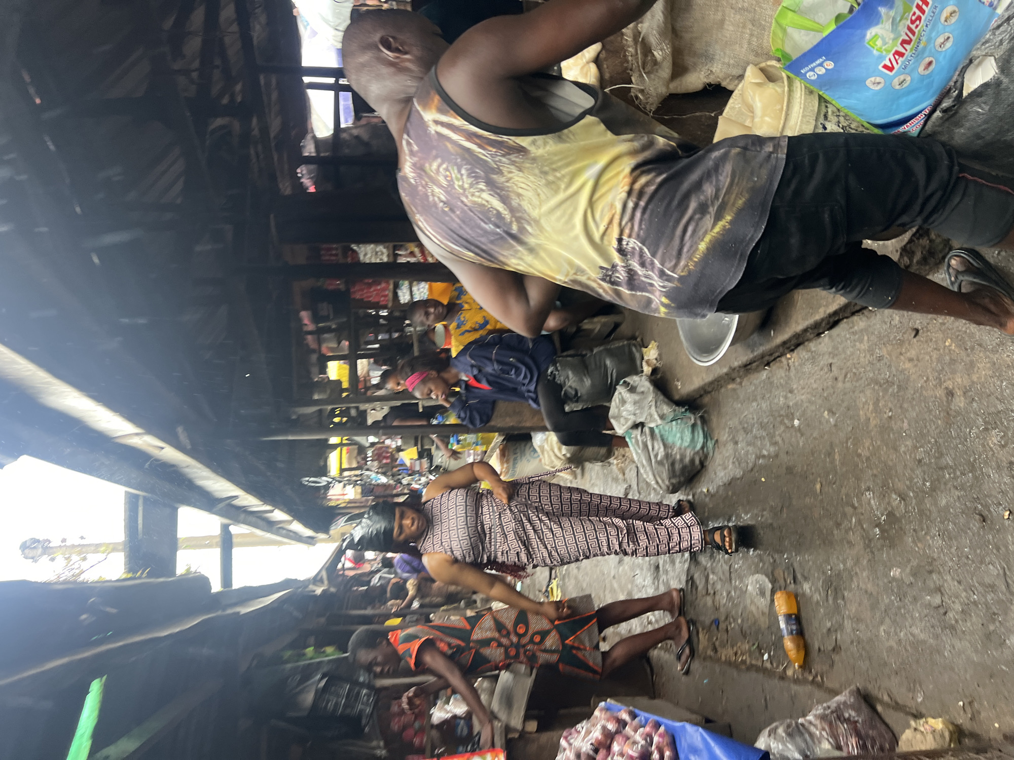 The image depicts a bustling marketplace scene in Nkpor, Nigeria, likely during rainfall given the wet ground and the presence of umbrellas or awnings overhead. In the foreground, a man wearing a tank top with a wolf graphic is seen from the back, facing away from the camera. To his left, a woman in a patterned jumpsuit walks past, her head covered with a black headscarf. Further in the background, other individuals are visible, some seated and others standing amidst the market stalls. The environment is characterized by the presence of goods displayed for sale, bags, and makeshift shelters, indicating a typical open-air market setting. The overall atmosphere appears active and somewhat chaotic, typical of a busy Nigerian market.