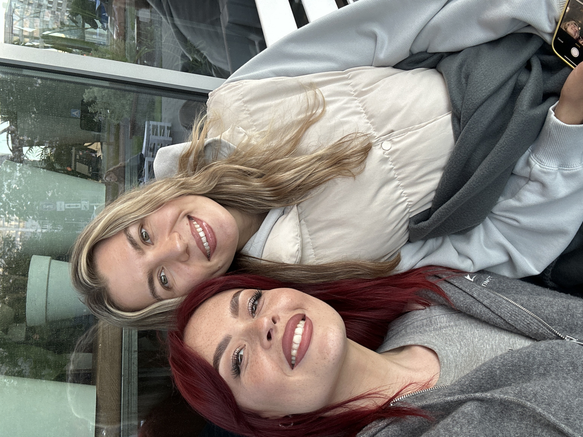 Two young women are captured in a close-up, rotated portrait, appearing to be lying down or leaning back together for a photograph. Both are smiling broadly at the camera, conveying happiness and contentment.
The woman in the foreground has vibrant red hair, freckles, and distinctive eye makeup. She is wearing a grey t-shirt layered under a grey zip-up hoodie or jacket, with the partial brand name "TING" visible on the chest of her jacket. The woman behind her has long, wavy blonde hair and is dressed in a light blue long-sleeved top and a light beige puffer vest. She holds a smartphone, likely an iPhone, near her head; its screen displays a camera interface with the words "Cancel" and "Photo" visible, along with a reflection of a face.
They are situated indoors next to a large window, which provides reflections of interior elements, such as a white chair or bench. Through the window, an outdoor scene is visible, featuring greenery and distant buildings, indicating a daytime setting. The overall mood is casual and friendly, suggesting a shared moment captured in Tolyatti, Russia.