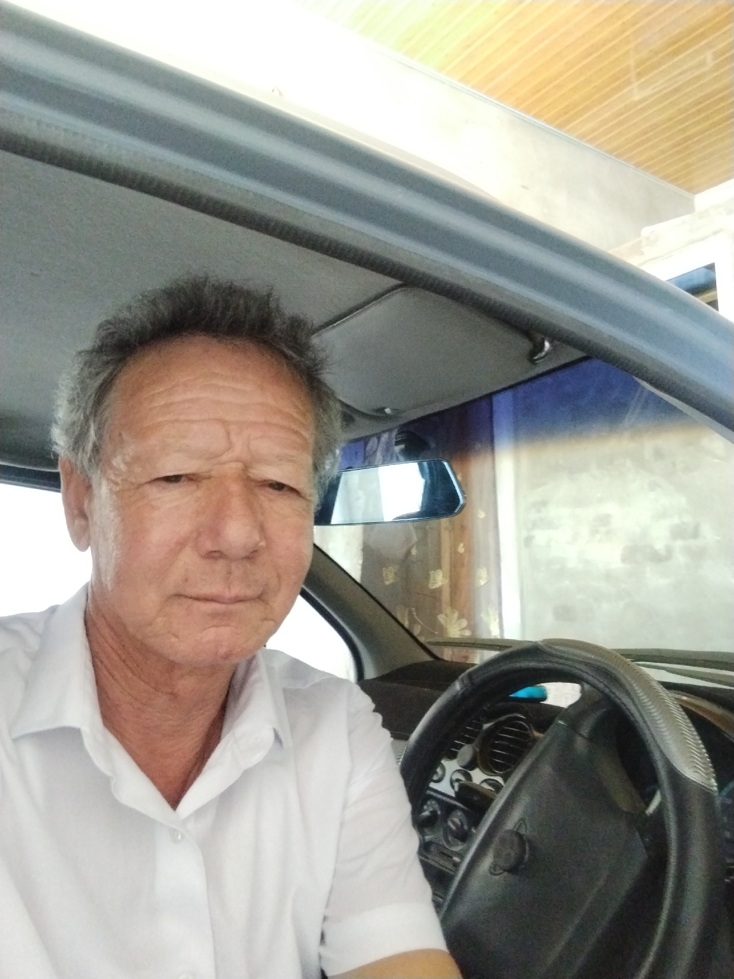 A middle-aged man with grey hair and wrinkles sits in the driver's seat of a car, looking at the camera. He is wearing a white button-down shirt. The interior of the car is visible, including the steering wheel and dashboard. The background appears to be a street scene in Shovot, Uzbekistan, with a building and trees.