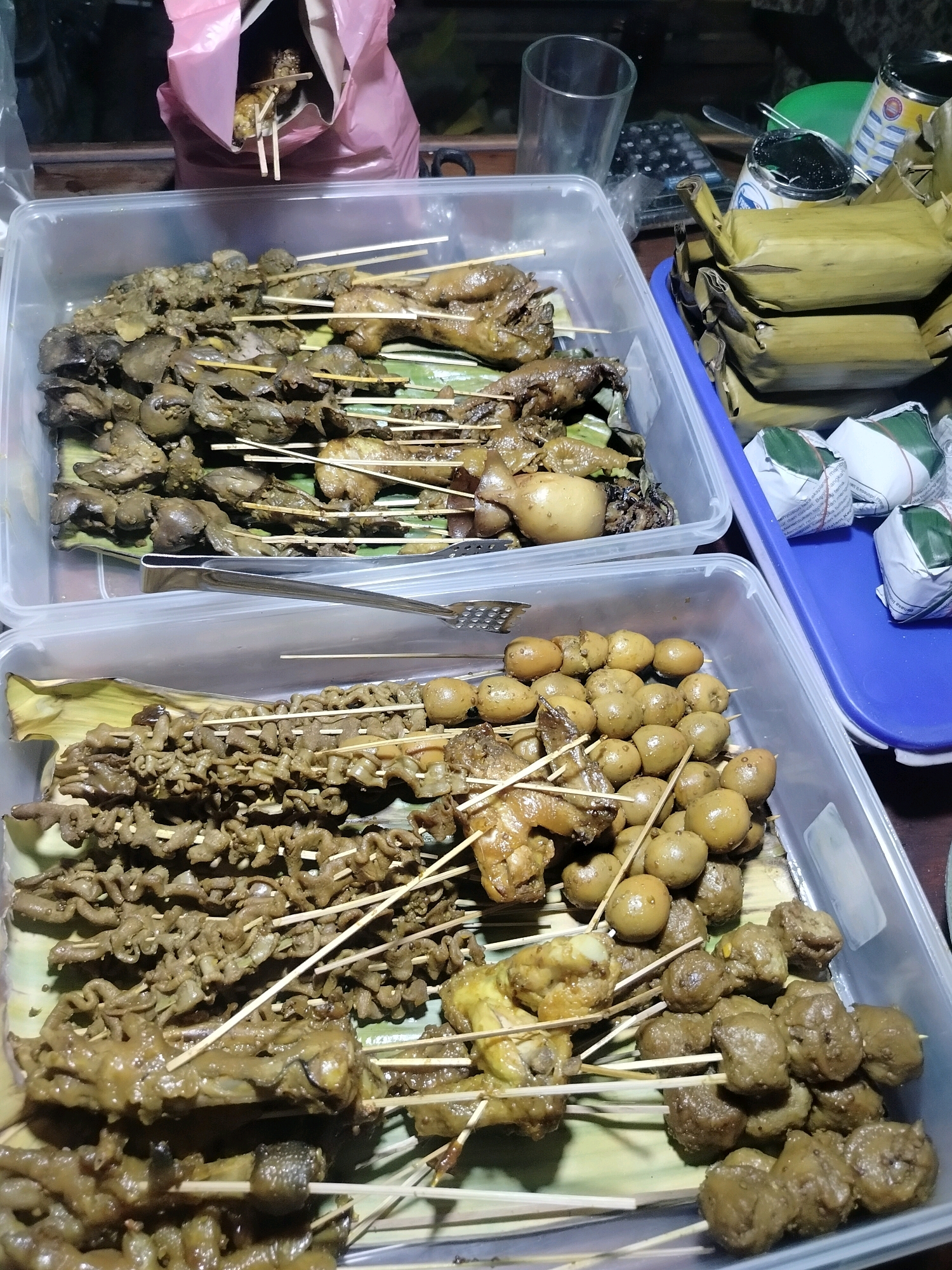 The image depicts an outdoor food stall in Kroya, Indonesia, likely at night. In the foreground, two clear plastic containers hold various Indonesian street food items on skewers. The top container features skewers of chicken parts and quail eggs. The bottom container displays skewers of intestines, chicken feet, and meatballs. The food is presented on banana leaves. To the right, a blue plastic tray contains wrapped food items, possibly rice or other snacks. In the background, a pink plastic bag is partially visible, and various objects like a calculator and a glass are on a surface, suggesting a busy vending environment. The lighting suggests it is evening or night.