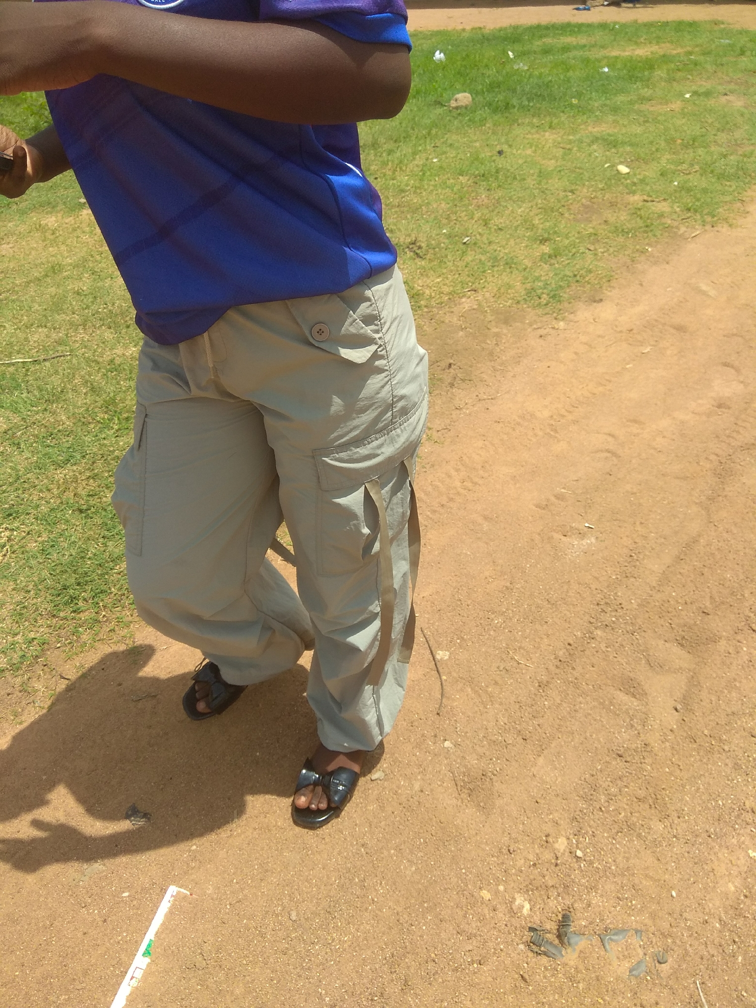In Jalingo, Nigeria, a person is seen from the waist down wearing a blue t-shirt and light grey cargo pants. The individual is also wearing dark colored sandals. The scene appears to be outdoors, with a grassy area to the left and a dirt path or ground on the right. The lighting suggests it is daytime.