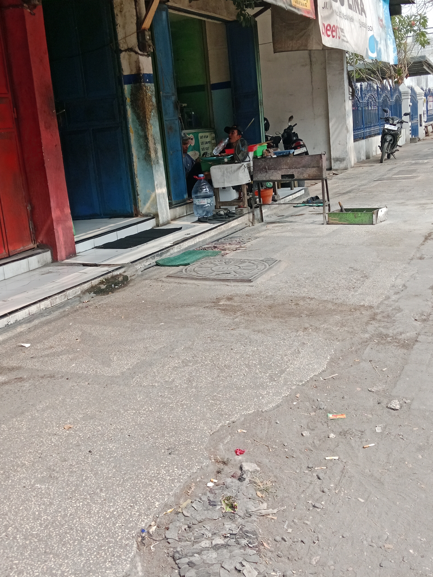 The image captures an outdoor scene in what appears to be a street or alleyway in Kediri City, Indonesia. In the foreground, the pavement is rough and uneven, with visible patches of gravel and dirt. There are scattered pieces of debris, including cigarette butts, small rocks, and what looks like a crumpled plastic wrapper. A circular metal manhole cover is set into the pavement.
To the left, a building has a red facade and blue doors. Adjacent to this, a blue doorway leads into a shop or eatery. Inside, two individuals are visible. One person, wearing a dark cap, is seated at a table and holding a bottle of water. Another person is also seated, partially obscured. A charcoal grill is set up in front of the establishment. A large plastic water bottle and several other items, including an orange bucket, are placed on the ground nearby. A motorcycle is parked in front of the building.
Further to the right, a blue metal fence lines the street, with another motorcycle parked near it. A white banner with text is stretched above the building's entrance. The overall environment suggests a commercial or semi-commercial area, likely a street-side food stall or shop. The lighting indicates it is daytime.