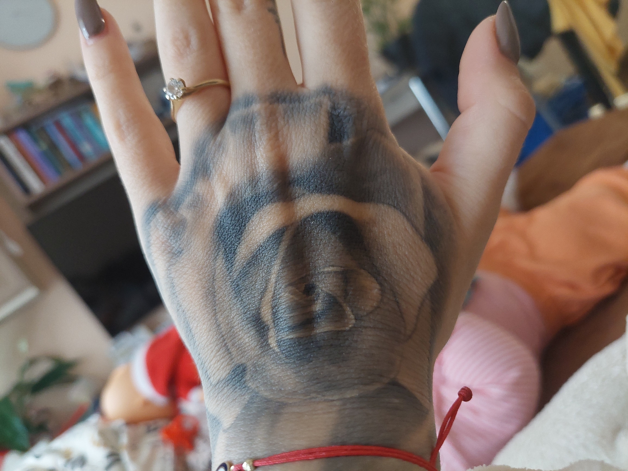 A close-up shot of a person's left hand with a large black and grey tattoo of a rose on the palm and the back of the hand. The hand has manicured nails with a taupe-colored polish. A gold ring with a clear stone is visible on the ring finger. A red string bracelet is worn around the wrist. The background includes a blurred image of a bookcase, a chair, a red and white object, and pink fabric. The location in the city of Plovdiv, Bulgaria, is not discernible from the image.