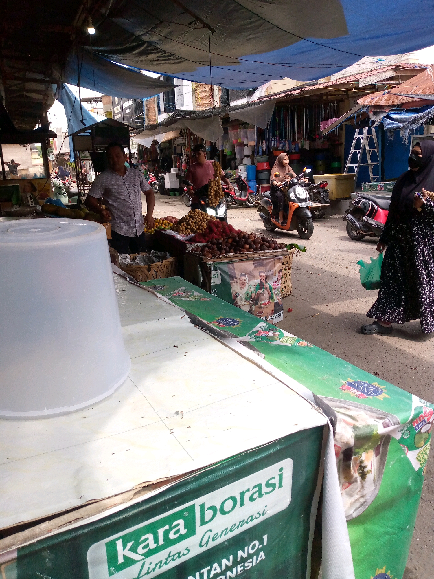 The image depicts a bustling marketplace scene in Dayahbaro, Indonesia. In the foreground, a large, translucent white plastic tub sits on a tiled surface. To the right, a green banner with white lettering reading "kara borasi" and "LINTAS GENERASI" is partially visible, extending to the right. Behind this, a woman in a black hijab and patterned black dress walks to the right, carrying a green plastic bag.
In the mid-ground, a man in a checkered shirt stands behind a display of fruits, including rambutan and other red fruits, in woven baskets. He appears to be interacting with a customer, though no specific action is clearly identifiable. Further back, a woman wearing a brown hijab rides a scooter with a child on the back, moving from left to right. Several motorcycles are parked in the background.
The marketplace is covered by a series of tarps, some blue and some translucent, creating shade. The stalls are filled with various goods, including what appear to be cleaning supplies like brooms and buckets. The overall environment suggests a typical outdoor market with activity and commerce. The architecture in the background indicates a mix of older buildings. The ground is paved with asphalt.