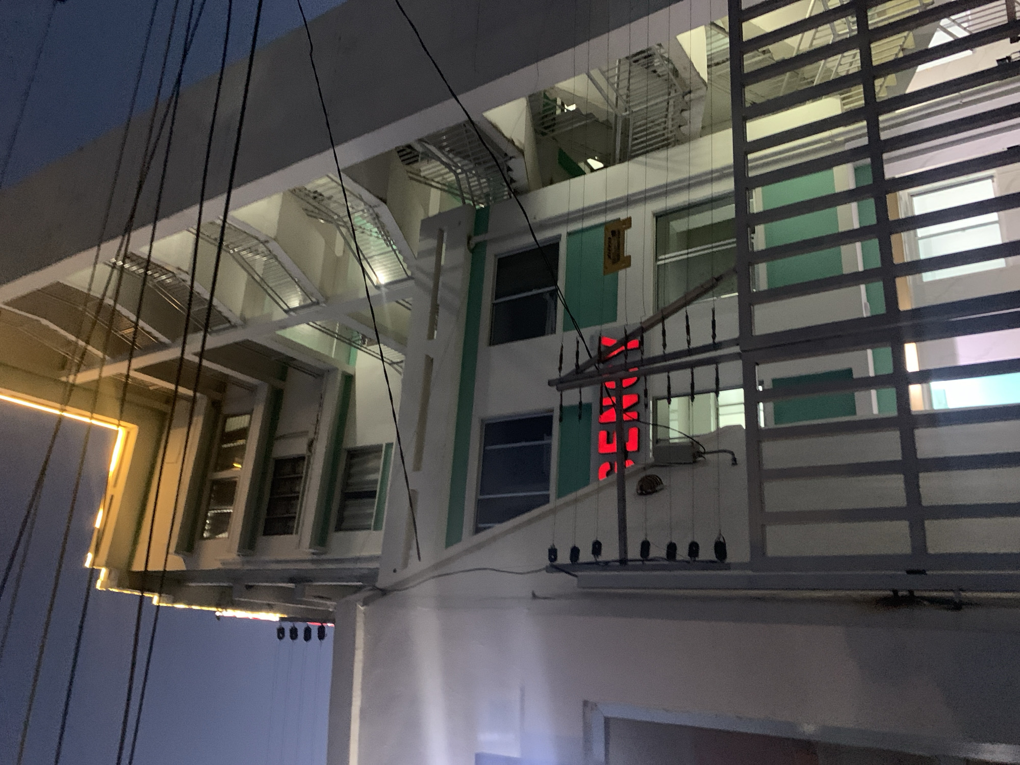 The image depicts a multi-story building in Dar es-Salaam, Tanzania, viewed from a low angle at night. The building features white walls with green accents and multiple windows. There are metal staircases visible on the upper floors, and a red neon sign is visible on the facade. A metal fence or gate is partially obscuring the view of the building. Electrical wires are strung across the foreground, further framing the building. The sky is a dark blue.