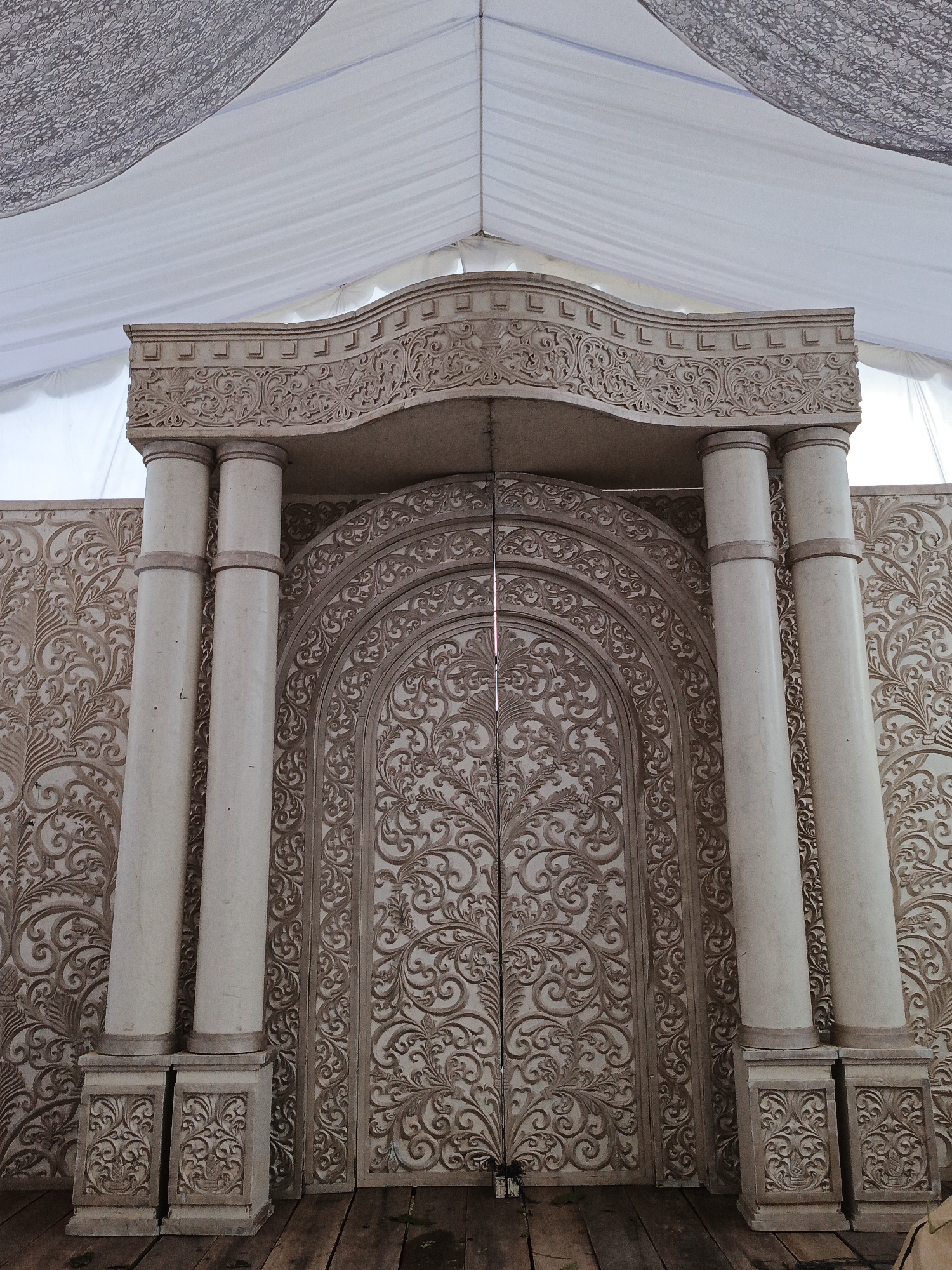 This image, captured in Sangso, Indonesia, depicts an elaborately decorated, temporary archway and double door structure. The central element is a light beige or off-white ornamental facade, featuring intricate baroque-style scrollwork, floral patterns, and geometric motifs across its entire surface. The archway's pediment has a curvilinear top edge, detailed with repetitive square relief elements and continuous carvings. Four slender, cylindrical columns, two on each side, support the pediment; these columns are a plain light color with two dark rings near their tops and rest on rectangular bases also adorned with carved scrollwork. The double door beneath the arch is topped with a matching curved outline and is entirely covered in dense, symmetrical carved patterns. Flanking the central arch are decorative panels extending outwards, which replicate the same ornate carving style. Overhead, a temporary white fabric tent structure is visible, with large swaths of white drapery. A section of lighter grey, possibly textured or lacy, fabric is layered over part of the white ceiling. The floor is composed of rough, dark wooden planks with visible grains and seams, exhibiting minor scuffs and a small piece of dark debris. The scene is evenly lit, indicating an indoor or covered outdoor setting.