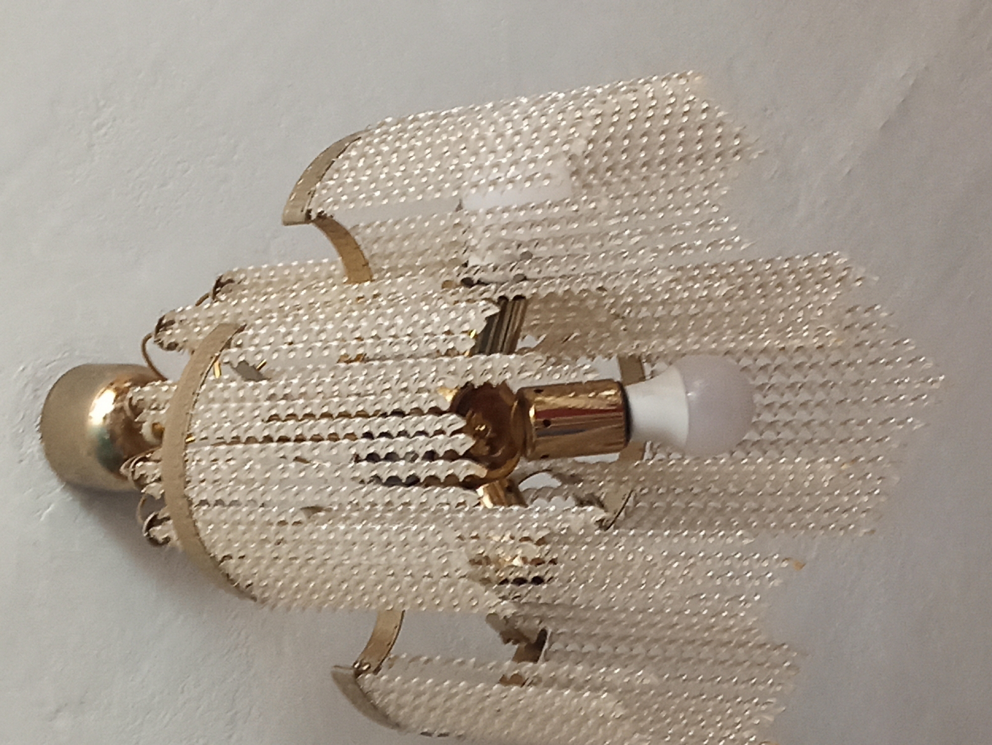 The image shows a close-up view of a vintage chandelier hanging from a textured white ceiling. The chandelier has a gold-colored metal frame with multiple tiers of clear, bead-like strands. A single white light bulb is visible, screwed into a gold-colored fixture at the center of the chandelier. The ceiling appears to be a rough, painted surface. The location in Masty, Belarus, is not discernible from the image.