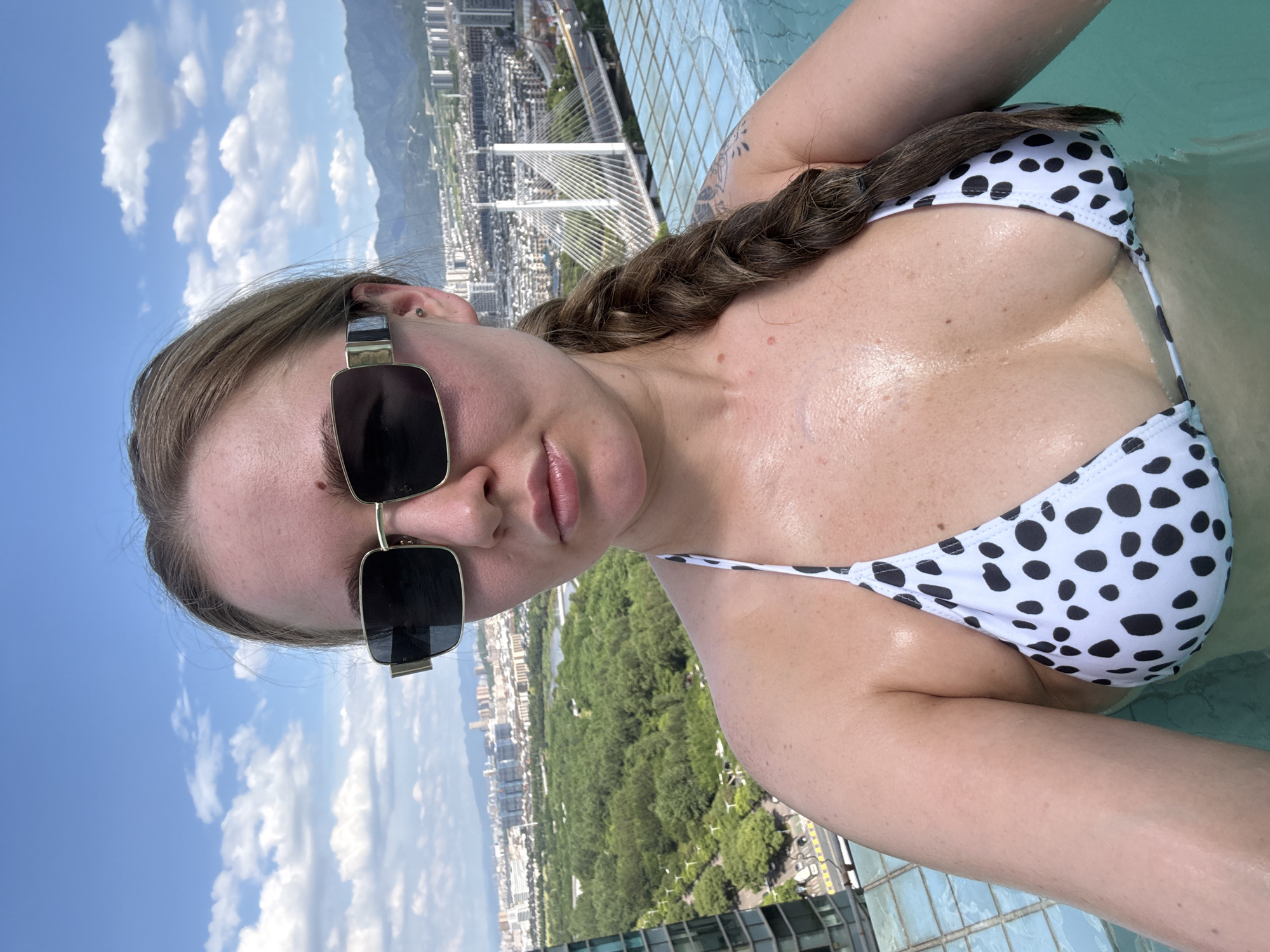 A woman with blonde hair braided over her right shoulder, wearing large, rectangular, gold-rimmed sunglasses and a white and black polka dot bikini top, is in an infinity pool on a sunny day. She is looking towards the camera with her mouth slightly open. Behind her, a city skyline with buildings and mountains under a cloudy blue sky is visible. This appears to be the rooftop infinity pool of a hotel in Yiwu, China, with a view of the city and the Yiwu International Trade City.