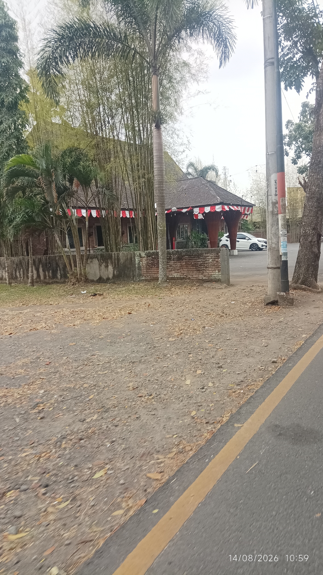 The image depicts a roadside view in Tasikmalaya, Indonesia, with a building featuring a distinctive roof and red and white decorative awnings visible in the background. The building appears to be constructed with brick and is surrounded by bamboo trees and palm trees. A white car is parked in front of the building. The foreground shows a gravelly area and a paved road with a yellow lane marker. A tall utility pole stands to the right of the building. The scene is captured on a cloudy day. The date and time stamp on the image indicates August 14, 2026, at 10:59.