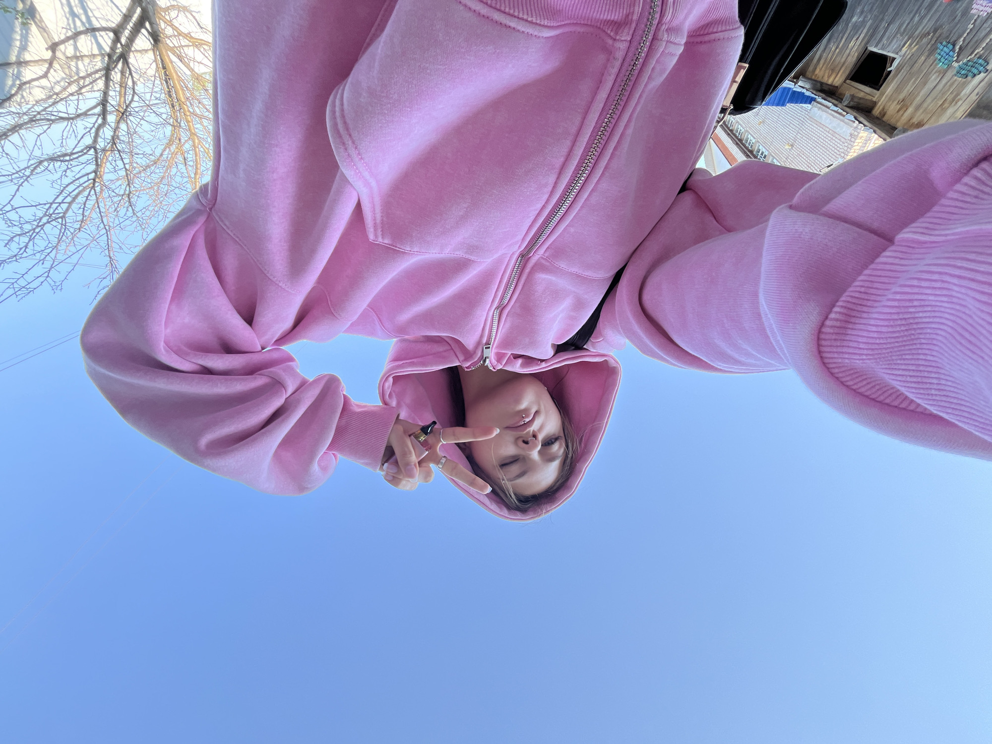 A young person wearing a pink hooded sweatshirt is looking down at the camera and making a peace sign. The sky is blue. There are tree branches and buildings in the background. The location is potentially in Фоки, Russia, based on the prompt's instruction.