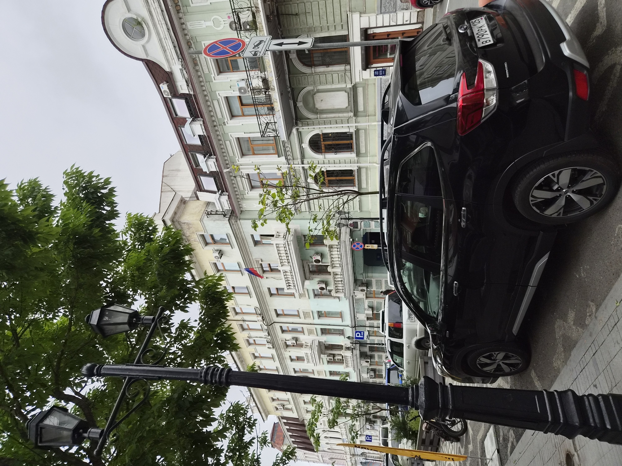 This is a street-level, eye-level, medium-shot photograph taken in Vladivostok, Russia, under an overcast sky. In the foreground, a black Subaru Forester is parked on the right side of the street. The vehicle's license plate is visible, reading "B790PK 125". A portion of a black metal lamppost is on the left side of the image. Behind the car, a row of multi-story buildings with light green and cream facades lines the street. The buildings feature classical architectural details, including balconies with white railings, arched windows, and decorative trim. A Russian flag is displayed on one of the buildings. Several other vehicles are parked along the street, including a white SUV. A person is visible walking in the distance. A parking sign and a no-standing sign are also present on the sidewalk. Trees with green foliage are visible in the foreground and between the buildings. The ground is paved with asphalt and some cobblestone.