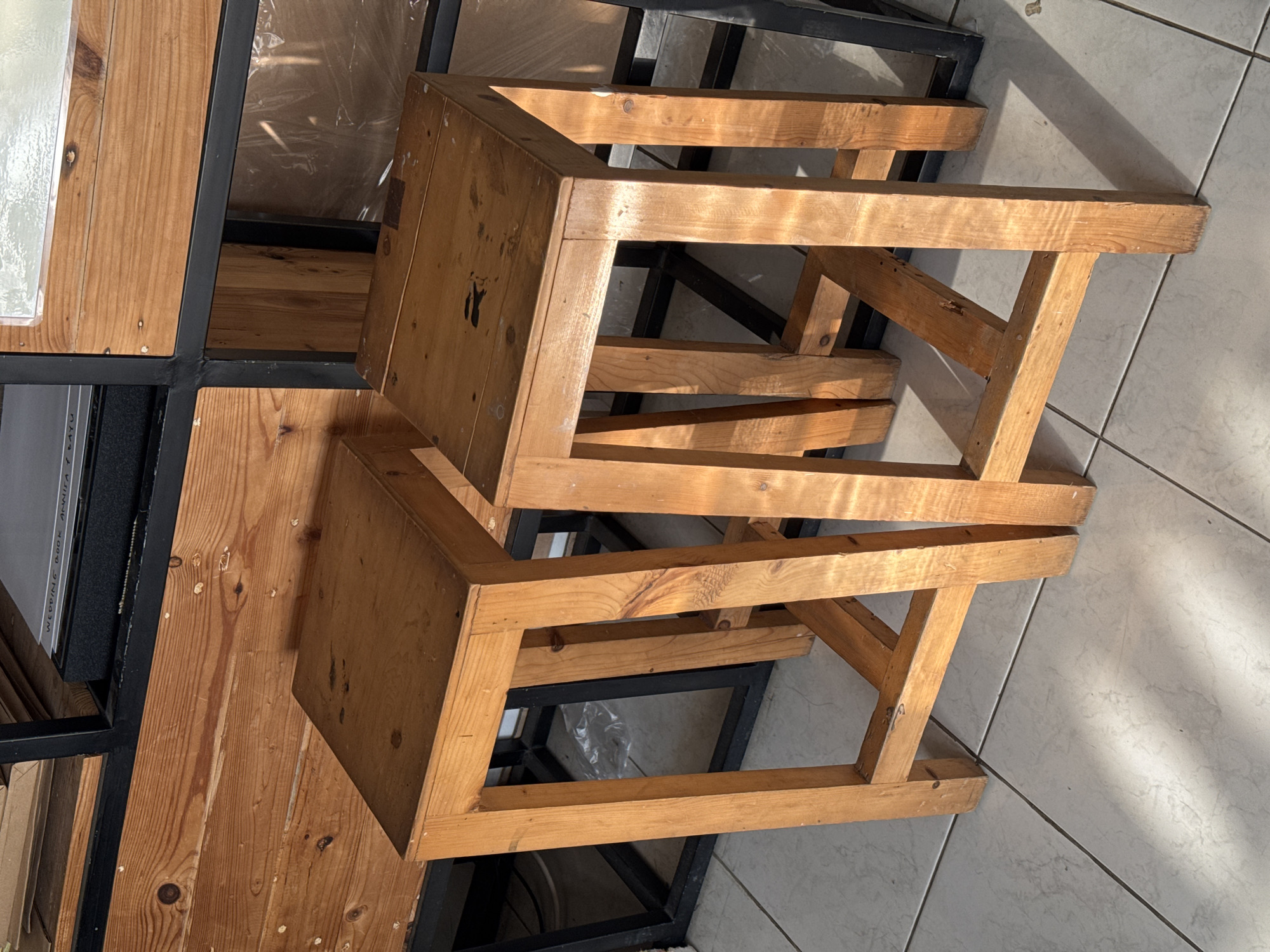 In Kedawung, Indonesia, two wooden stools stand on a tiled floor bathed in sunlight. The stools are simple, with square seats and four legs connected by crossbars. Behind the stools, a wooden shelf unit with black metal framing is visible, displaying various items on its shelves. The overall scene suggests an indoor setting, possibly a shop or a workshop, with a focus on the rustic charm of the wooden furniture.
