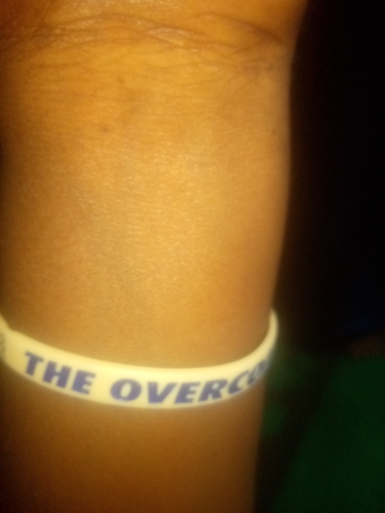 A white wristband with blue text is visible on a person's arm. The text appears to read "THE OVERCO". The arm is a light brown color, and the background is dark, with a hint of green. The image is a close-up shot, with the wristband being the primary focus. The setting is not identifiable as being in Jauro Yinu, Nigeria.