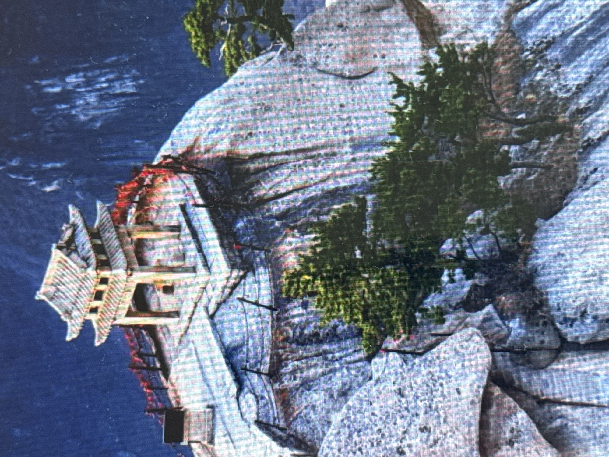 The image shows a traditional Japanese-style building, possibly a pagoda or temple, perched precariously on the edge of a steep, rocky cliff. The building appears to be made of wood and has multiple tiers, with curved roofs and intricate detailing. It is painted in vibrant colors, including red and gold, which stand out against the natural backdrop.
The setting is a natural landscape with a body of water below the cliff, likely a river or lake, indicated by the dark blue color and some lighter streaks suggesting ripples or reflections. The cliff face is rough and textured, with various shades of gray and white rock. Green foliage, likely trees or bushes, grows on the cliff and around the building, adding a touch of life to the scene.
The overall impression is one of dramatic contrast between the man-made structure and the rugged natural environment. The building seems to defy gravity, clinging to the cliff face. The image quality is somewhat grainy, suggesting it might be a photograph of a screen or a low-resolution digital image. There is no indication of people or specific activities taking place. The lighting suggests it could be daytime, but without further context, the time of day and weather are difficult to determine. The location cues do not specifically point to Pushkino, Russia, but rather to a mountainous or cliffside landscape that might be found in East Asia, given the architectural style of the building.