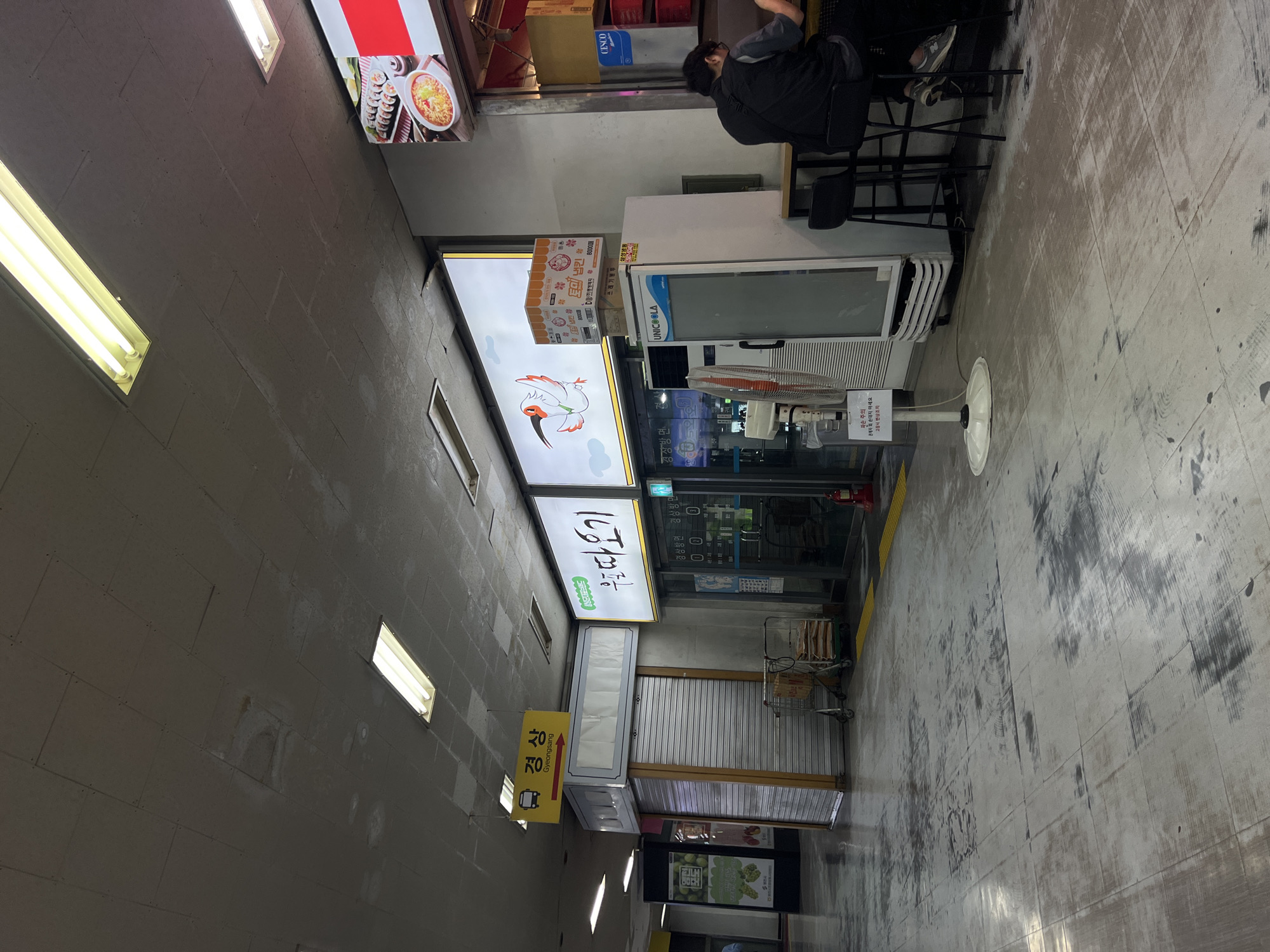 The image shows an outdoor alleyway or walkway between buildings, likely in Seoul, South Korea, as indicated by the Korean text. The walkway has a gray, textured floor that appears somewhat worn. On the left side, there are concrete block walls with rectangular fluorescent lights.
On the right side of the walkway, there are several signs and storefronts. One prominent sign has Korean text and an illustration of a bird. Below this, another sign points in a direction labeled "경상 Gyeongsang" with an arrow and a bus icon, suggesting it's a directional sign for a bus stop or transportation hub. Further down, there's a shop with a glass door and shutters. A shopping cart is visible outside this shop.
A person is partially visible in the upper right corner, sitting on a stool and facing away from the camera, appearing to be at a counter or table. There are some boxes and a refrigerated display unit (labeled "UNICOOLA") behind the person. Another sign below the "우도따오기" sign has Korean text that appears to be product information, mentioning "80000円", which is Japanese Yen, suggesting possible cross-cultural commercial activity or influence. A sign near the shuttered shop reads "파손 주의 관계자 외 손대지 마세요 고장시 변상조치" which translates to "Caution: Damaged. Do not touch except by authorized personnel. Compensation will be made in case of malfunction." This indicates a caution notice for equipment or merchandise.
The lighting suggests it is daytime, with the fluorescent lights casting a bright glow. The overall atmosphere appears to be that of a commercial or service area, possibly a market street or a row of shops.