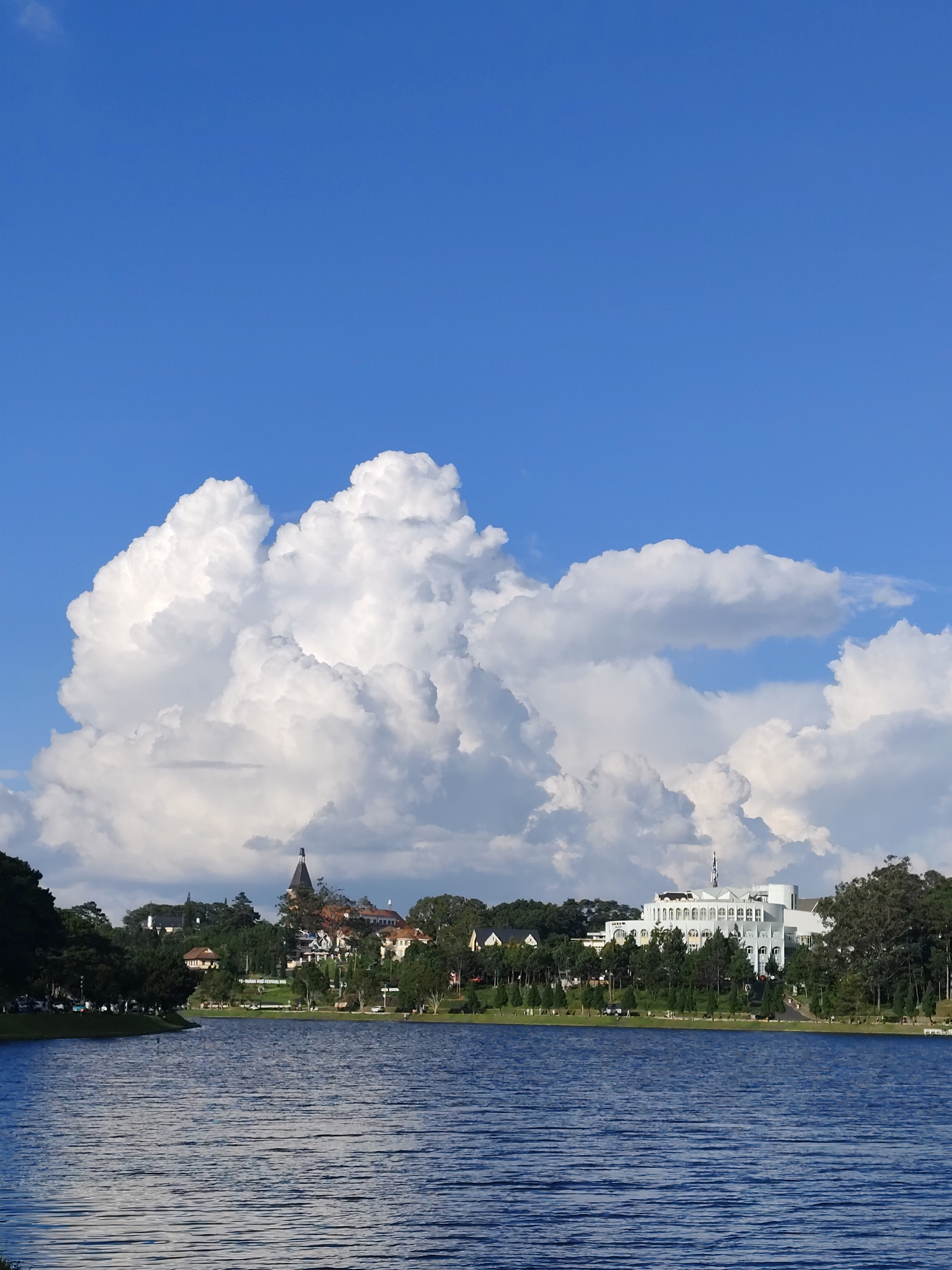 The image depicts a serene waterfront scene in Xuan Huong Lake, Da Lat, Vietnam. The foreground is dominated by the calm, blue water of the lake, reflecting the bright, clear sky and the surrounding scenery. The water exhibits subtle ripples, indicating a gentle breeze. Along the far bank of the lake, a line of lush green trees forms a natural border. Behind the trees, several buildings are visible. On the left, a cluster of buildings with reddish-brown roofs are nestled amongst the trees, with a distinctive building featuring a dark, pointed spire. Further to the right, a large, white, multi-story building with arched windows and a symmetrical facade stands out. The sky above is a vibrant blue, with dramatic, puffy white cumulus clouds congregating in the upper portion of the image, casting soft shadows on the landscape below. The overall impression is one of tranquility and natural beauty, with a touch of architectural charm from the distant buildings.