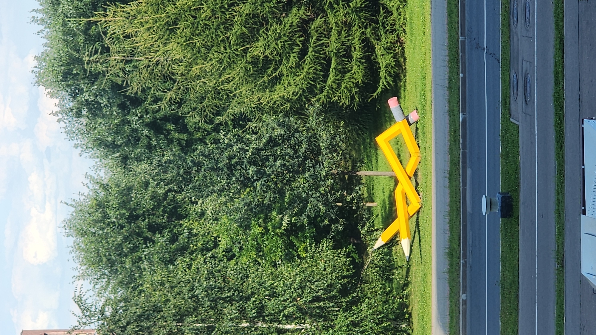 An outdoor scene depicts a large, yellow, stylized pencil sculpture lying on the grass. The sculpture is bent and appears to be made of metal or plastic. One end of the pencil has a white, pointed tip, and the other end has a pink eraser. The sculpture is situated in front of a dense line of green trees and bushes, with a portion of a tall evergreen tree visible on the right. In the foreground, there is a paved road with white lane markings, and a sidewalk area with a small, white-capped cylindrical structure on a dark rectangular base. The sky above is partly cloudy with white clouds against a light blue background. The overall impression is of a public art installation in a park-like setting within a city.