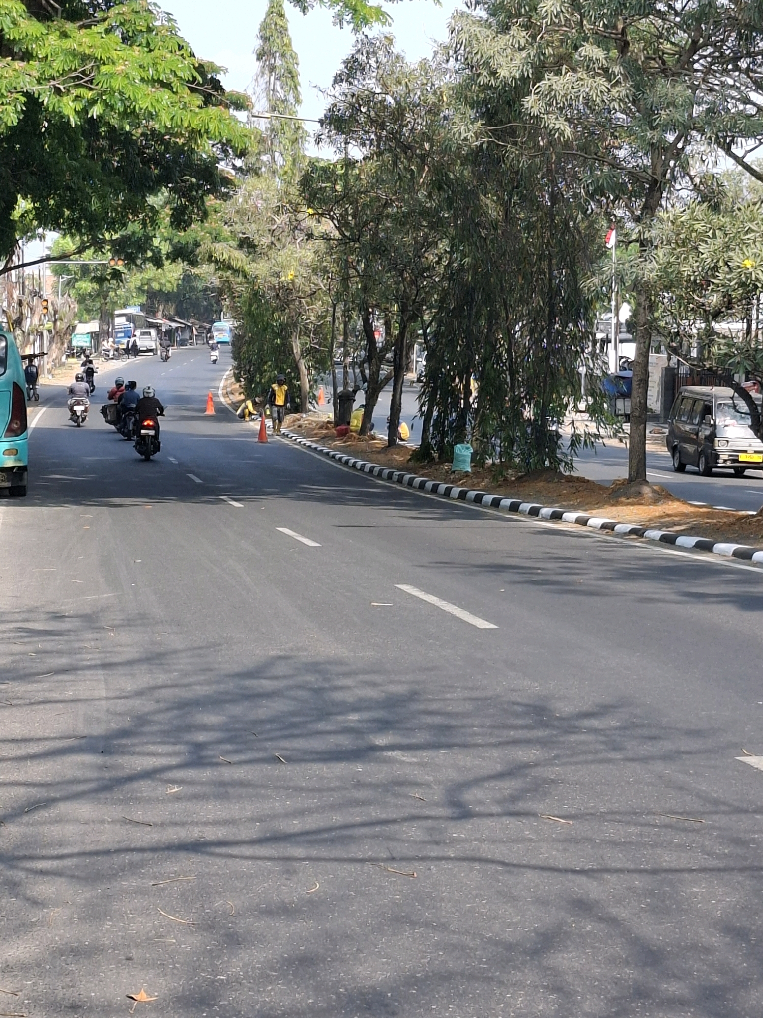 The image depicts a street scene in Sukabumi, Indonesia, on a clear, sunny day. The road, marked with white dashed lines, is asphalt and shows some debris. A line of trees with lush green foliage flanks the right side of the road, with some trees also visible on the left, casting shadows on the asphalt. The sky is bright and clear.
On the road, several individuals are riding motorcycles. In the foreground and mid-ground, there are at least five motorcycles with riders. One motorcycle in the mid-ground has a pillion rider. Further down the road, more vehicles, including a light-colored van on the right and what appears to be a turquoise bus on the far left, are visible. Several traffic cones are placed along the right side of the road, some orange and some possibly yellow. A person in a yellow jacket is standing near some of the traffic cones on the right shoulder.
The environment suggests an urban setting with sidewalks and roadside vegetation. The buildings visible in the background are modest, with some appearing to be shopfronts or small establishments. A flagpole with an Indonesian flag is partially visible among the trees on the right. The overall impression is of a moderately busy street in Indonesia during daylight hours.