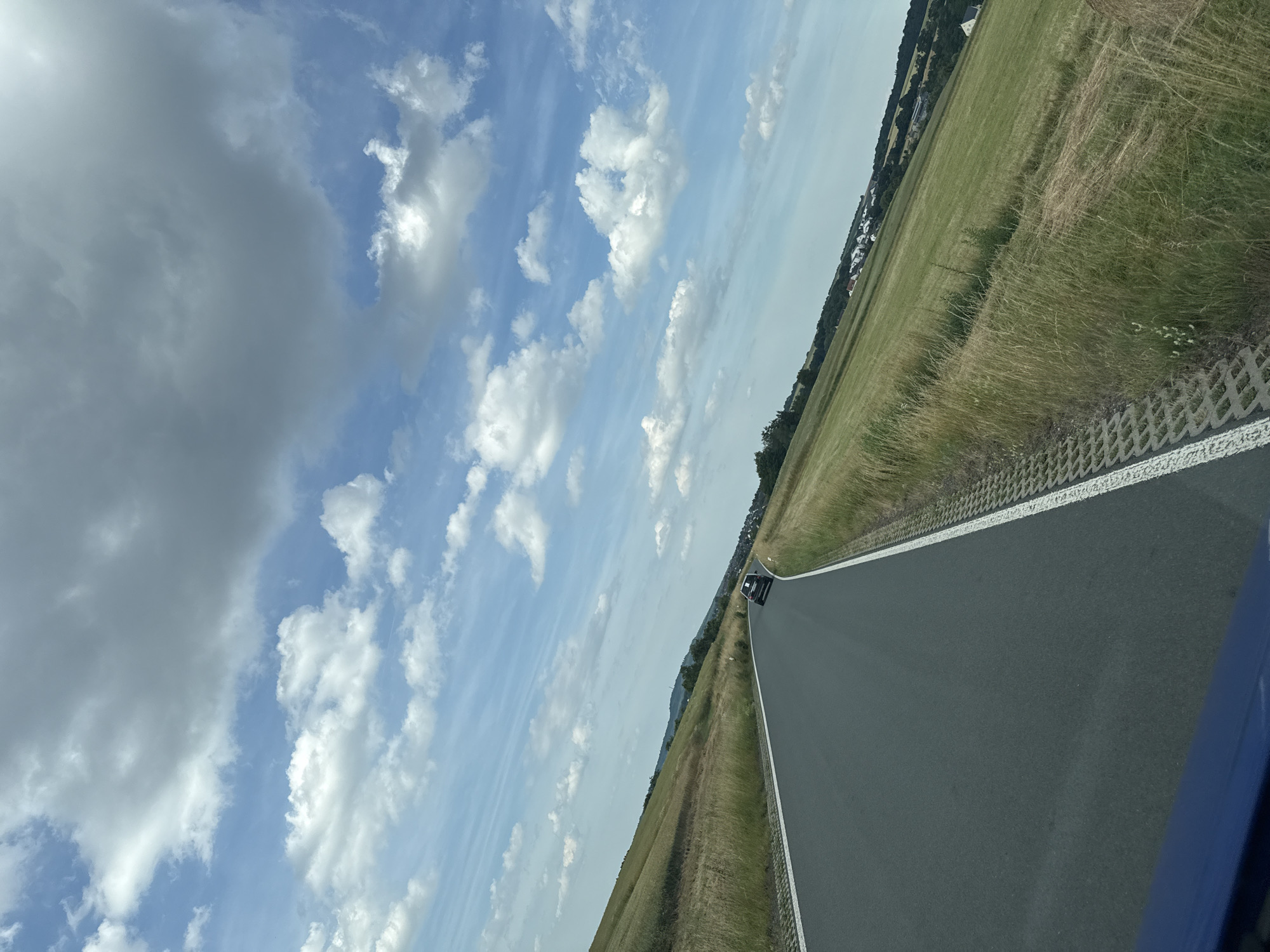 A black car drives on a grey asphalt road through a rural landscape with green fields under a cloudy blue sky. The road curves to the left, bordered by a white line and a gravel strip. The fields are covered in grass and wildflowers, sloping upwards and away from the road. In the distance, a village with white houses is visible nestled among trees. The image was taken in Niederwetz, Germany.