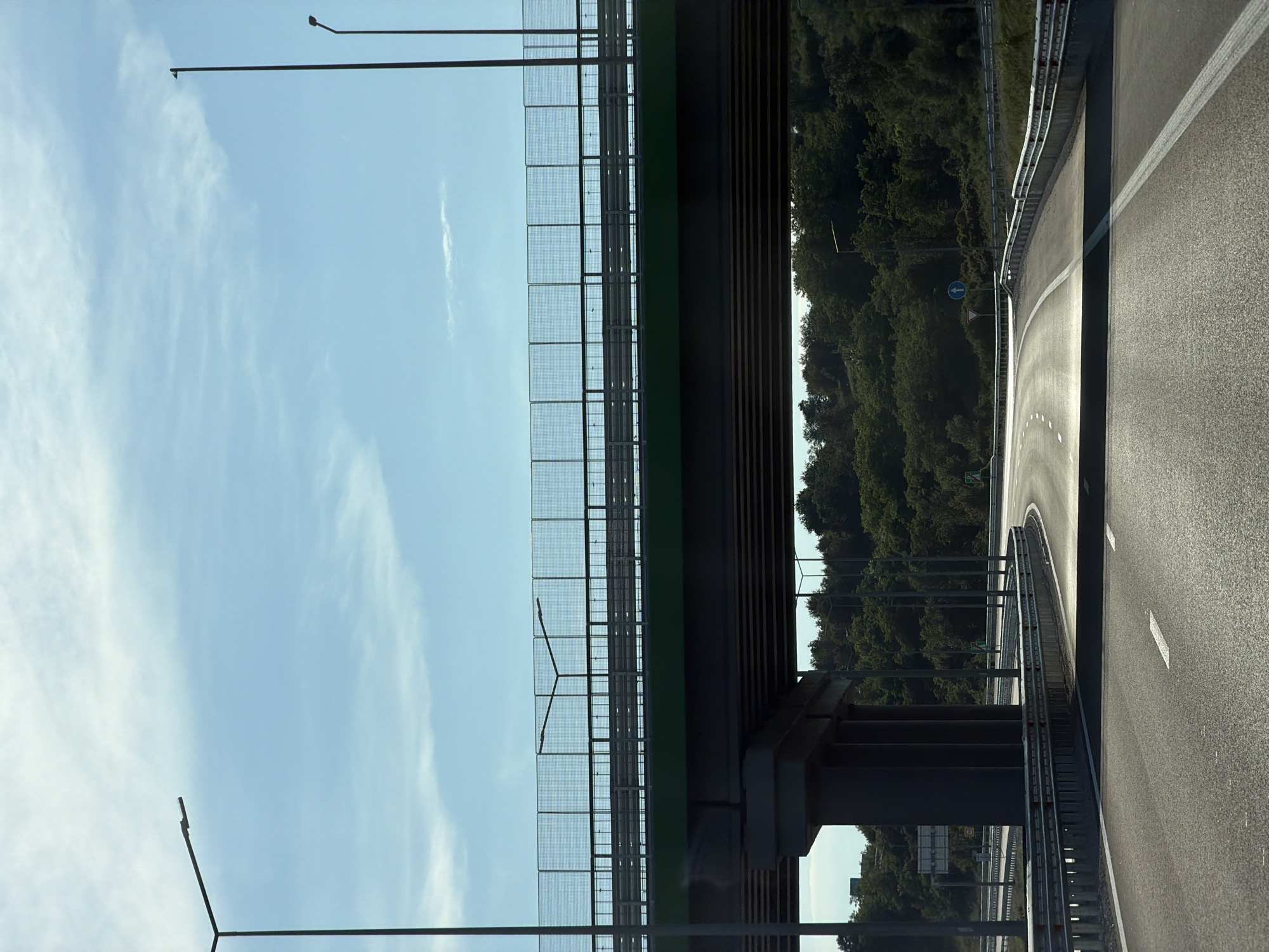 The image shows a highway overpass in Brestnitsa, Bulgaria, on a clear, sunny day. The overpass, painted green underneath and with a wire mesh barrier on top, casts a shadow over the road below. The road itself is asphalt with white lane markings. Several streetlights are visible along the sides of the highway. The surrounding area is covered with dense green trees, indicating a rural or semi-rural setting. The sky is a light blue with scattered wispy clouds. There are no people or vehicles visible in the frame, suggesting an empty stretch of highway. Road signs are present, including a blue circular sign with a white arrow pointing upwards, indicating a mandatory direction.